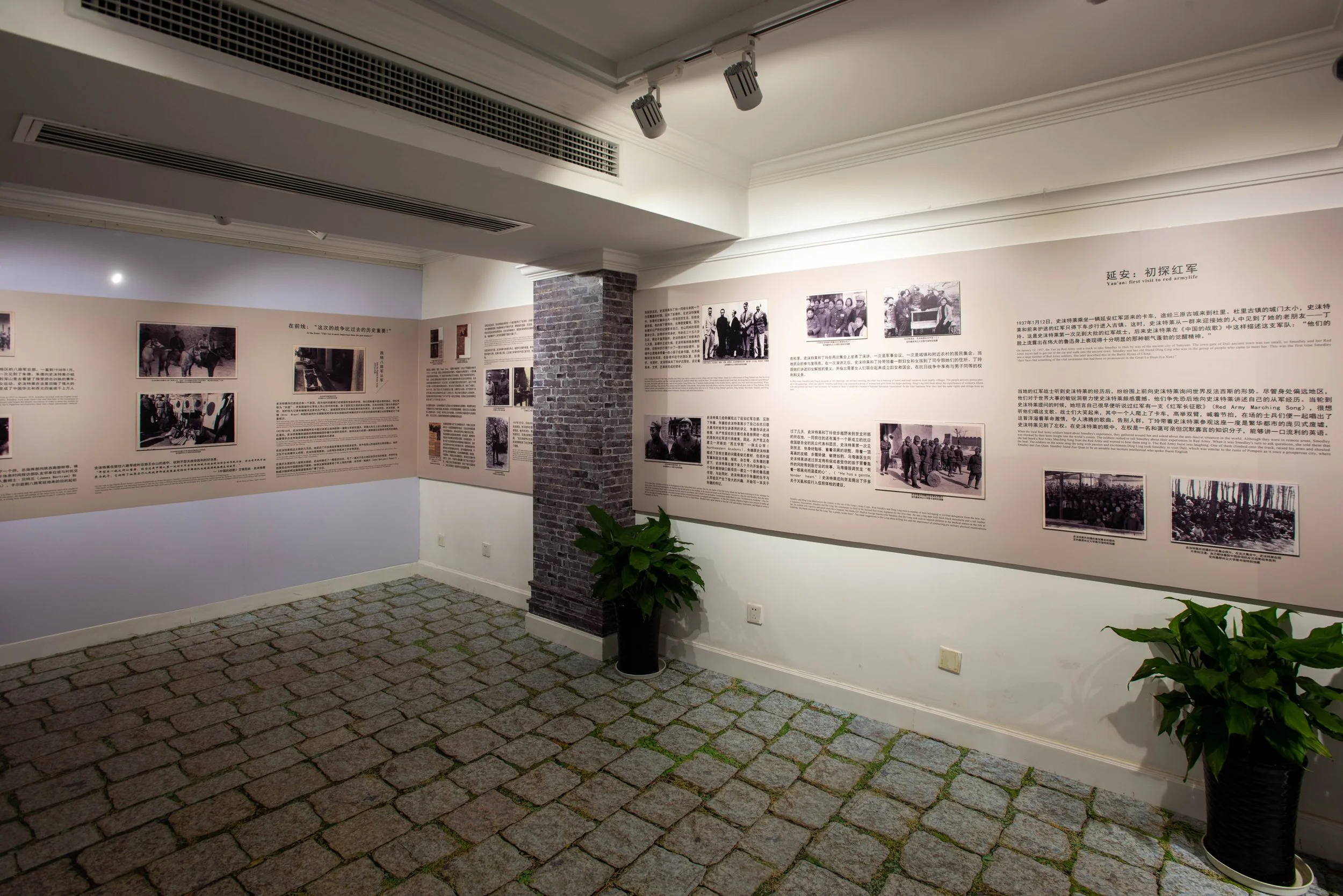 An indoor exhibition space with beige walls covered in historical photographs and Chinese text. The room features a brick pillar, green potted plants, and a stone-tiled floor. Track lighting is installed on the ceiling overhead.