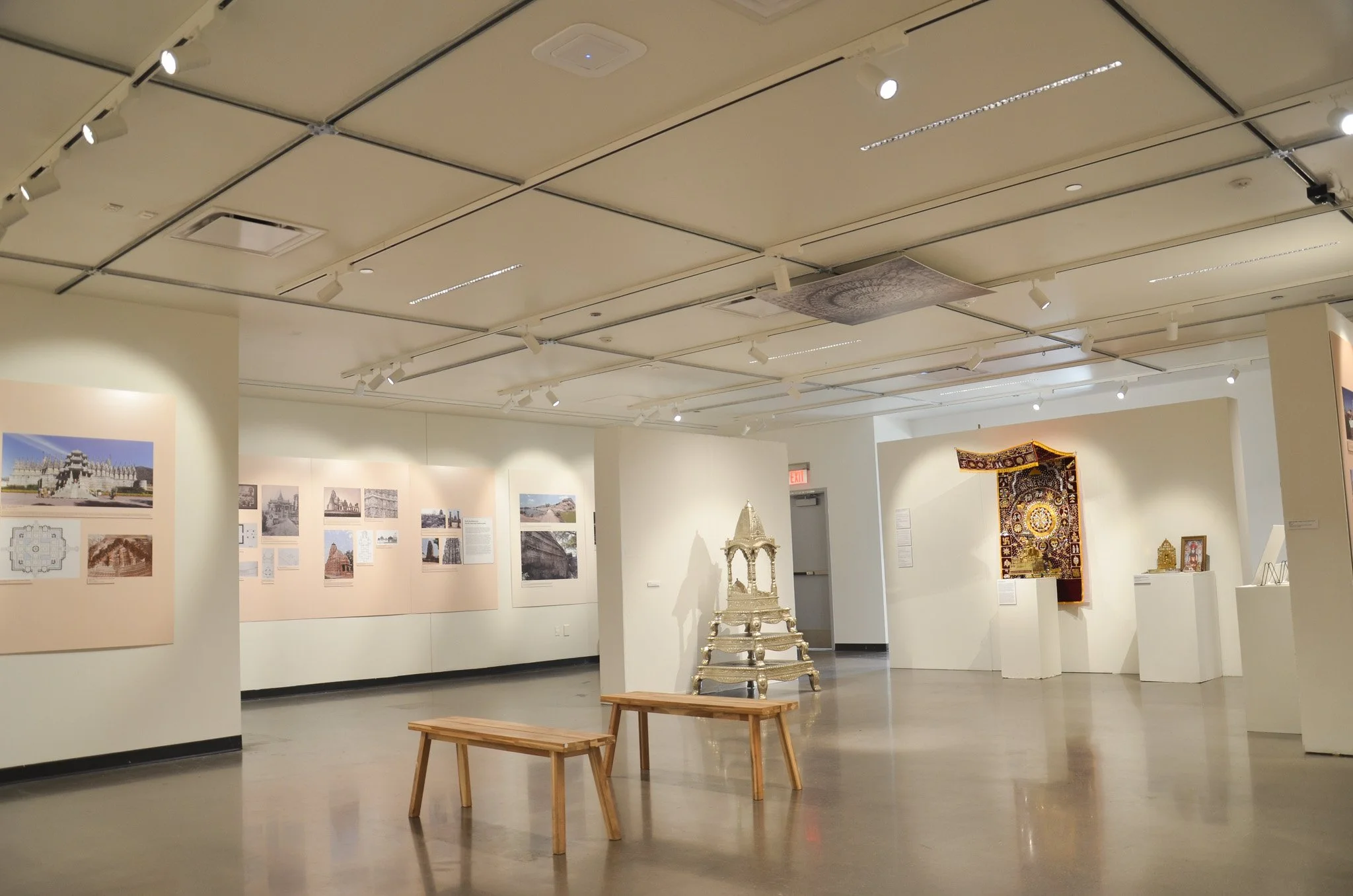 An art gallery with white walls displaying photographs of temples and buildings on the left, with sculptures, a tapestry, and benches in the foreground, under a ceiling with track lighting.