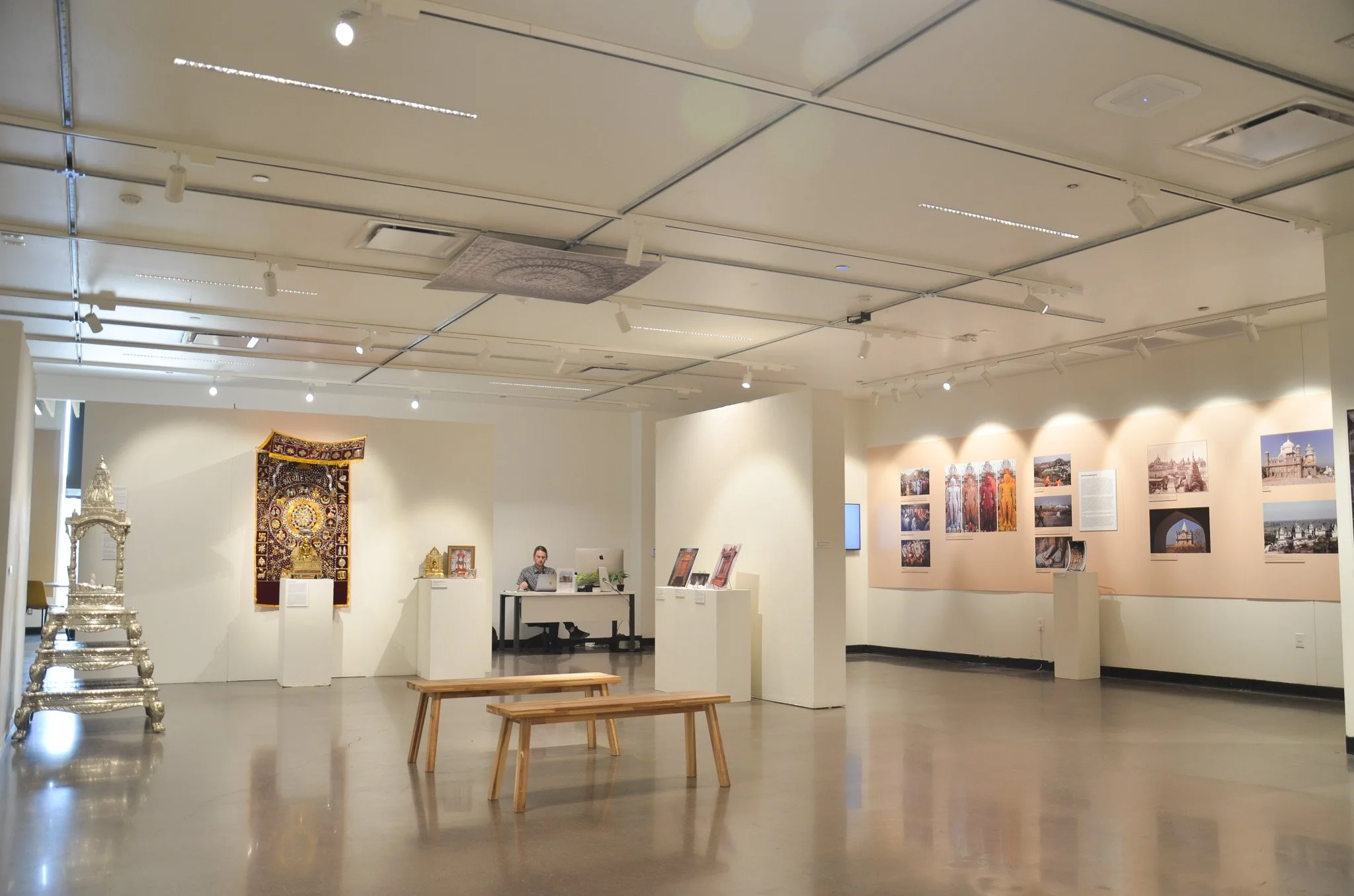 An art gallery interior with wall-mounted photographs, a person working at a desk, benches, and sculptures, all under a white ceiling with track lighting.