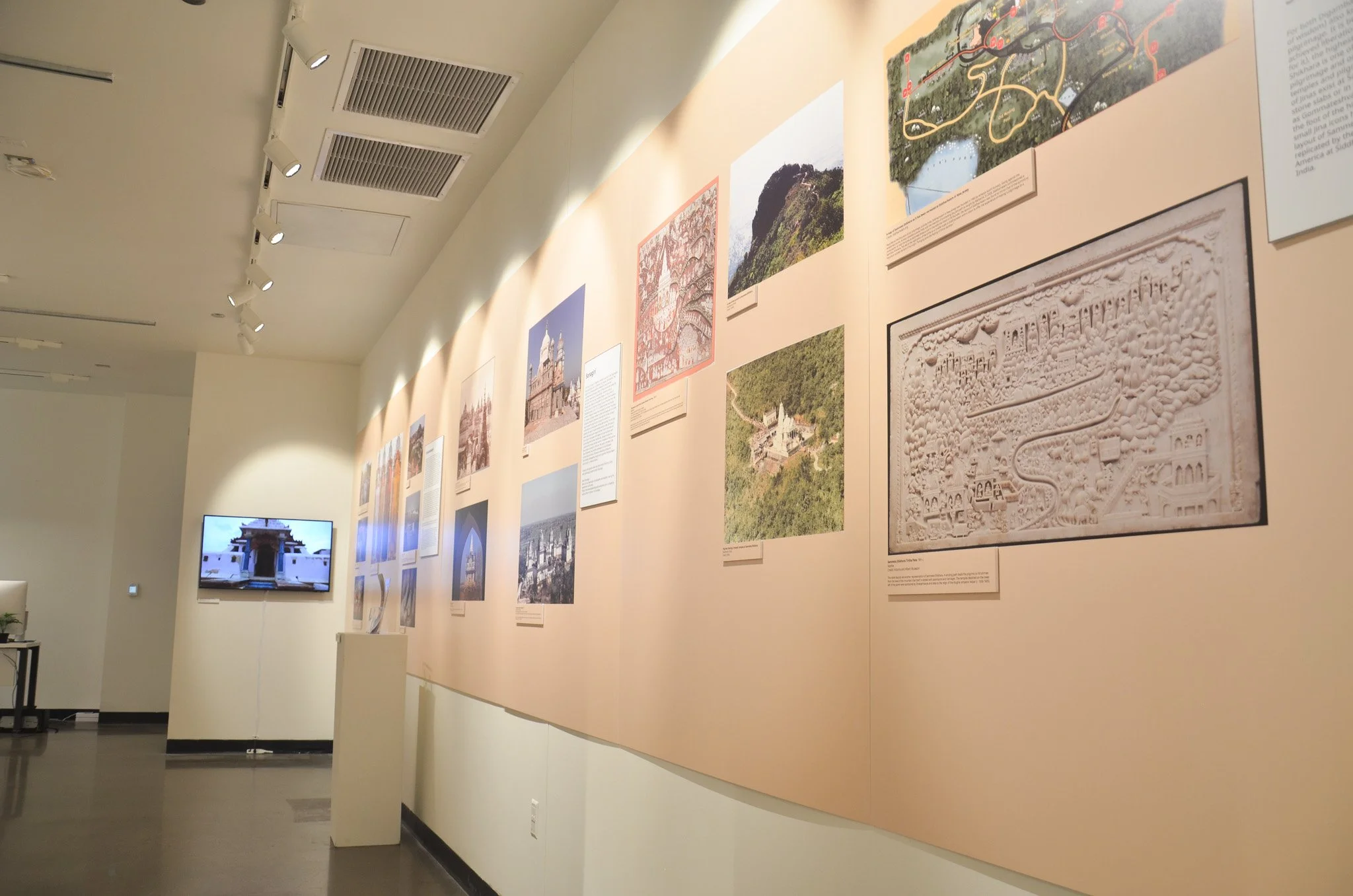 Gallery exhibit with framed photographs, descriptions, and a video monitor on a neutral wall in an art museum or gallery.