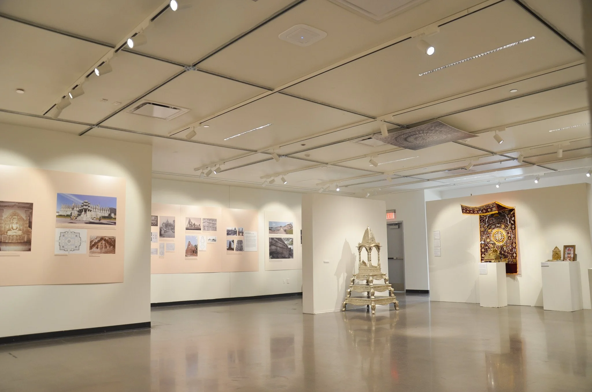 Interior view of an art gallery with beige and white walls, displaying photographs and artworks. A decorative silver throne is in the center, with a tapestry hanging on one wall. Track lighting illuminates the space, and an exit sign is visible in th