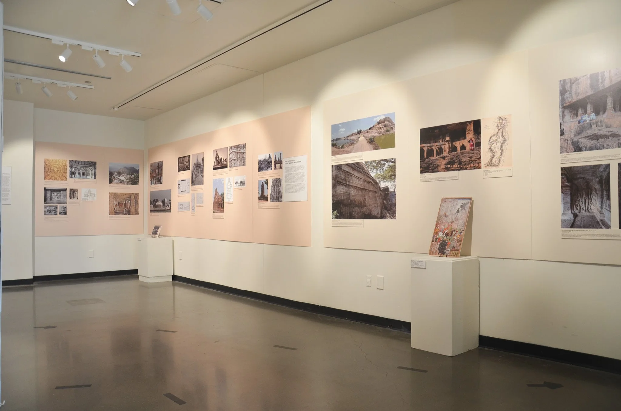 An art exhibit with photographs and images displayed on cream-colored walls in a gallery space, with a light-colored polished floor and track lighting on the ceiling.