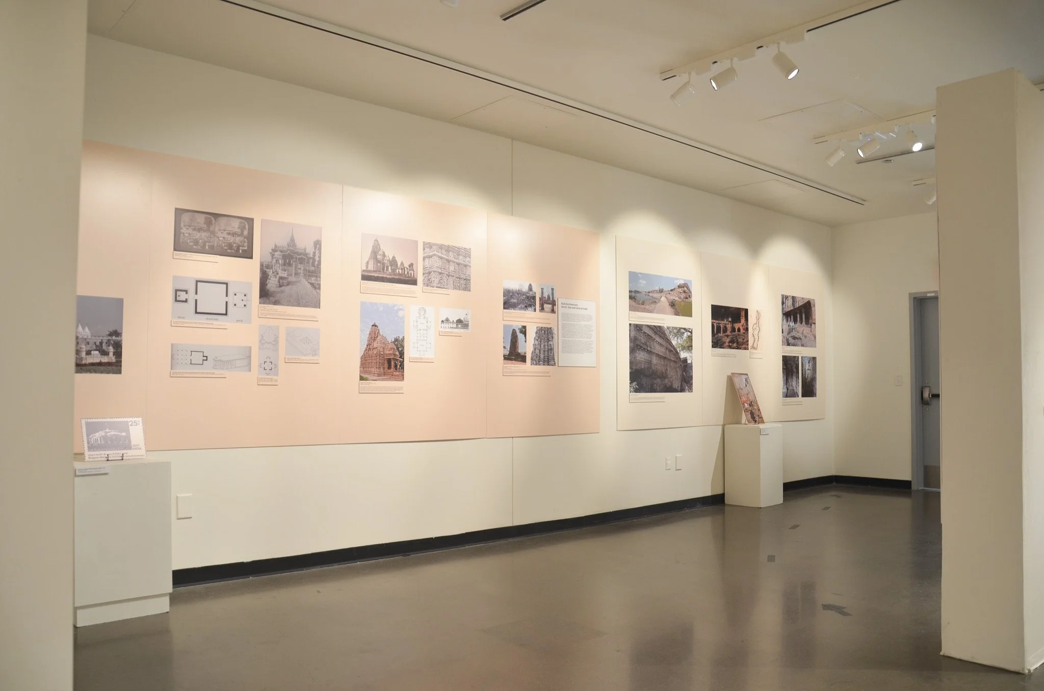 An indoor art gallery with a beige wall displaying black and white photographs and prints, including images of temples and ancient structures, and a white pedestal in front. The ceiling has track lighting illuminating the artwork.