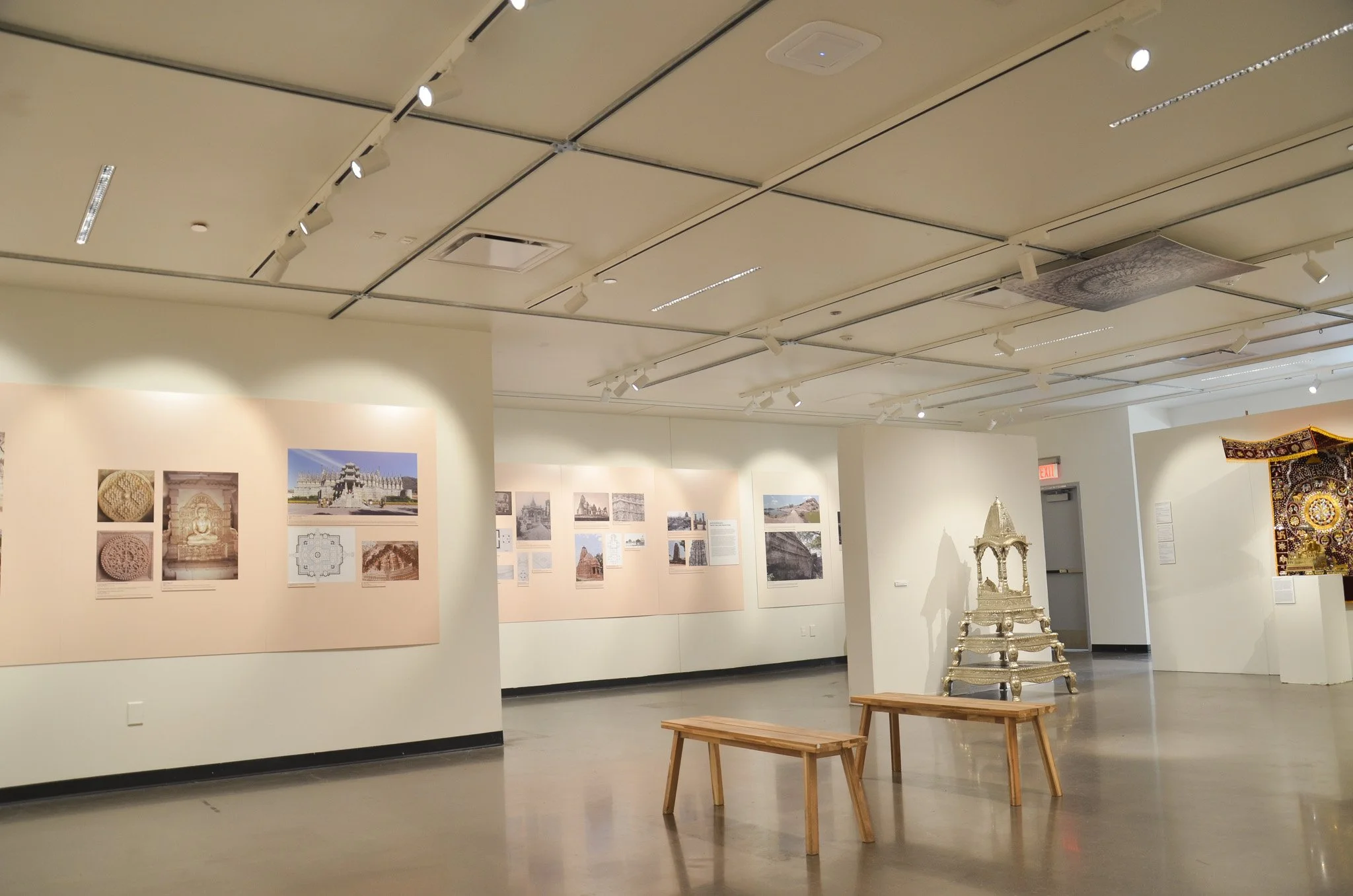 Interior of an art gallery with framed photographs and artworks on the walls, a carved silver throne, wooden benches, and ceiling lights.