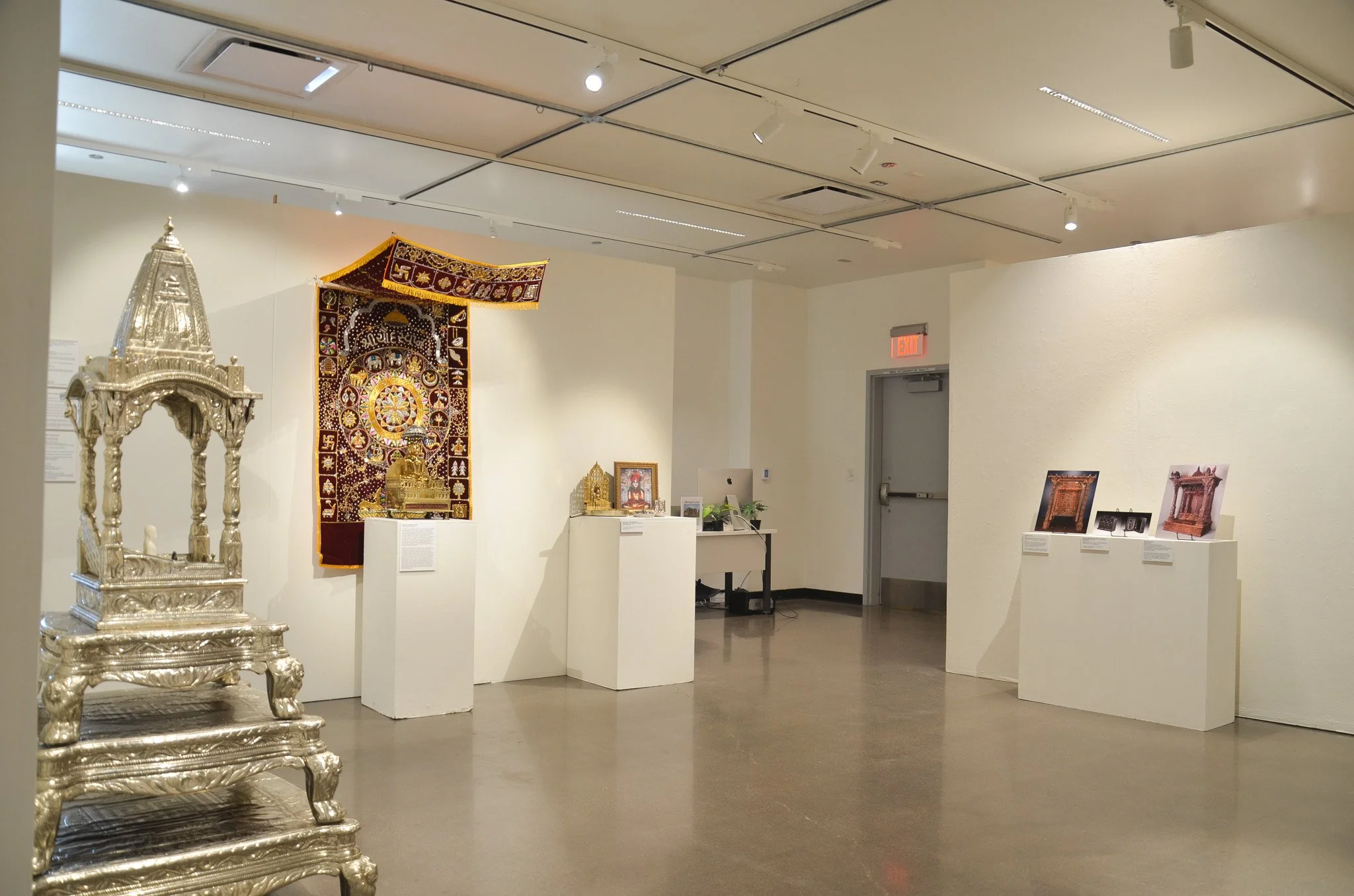 Museum exhibit showcasing ornate artifacts, including silver sculptures and decorative textiles, on white display stands in a well-lit room with white walls and ceiling.