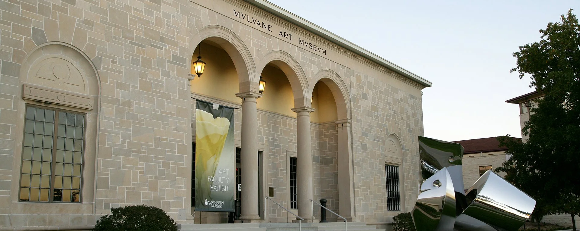 Exterior of the MvLune Art Museum with an Art Faculty Exhibit banner, arched windows, and a modern sculpture in the front yard, during daytime.