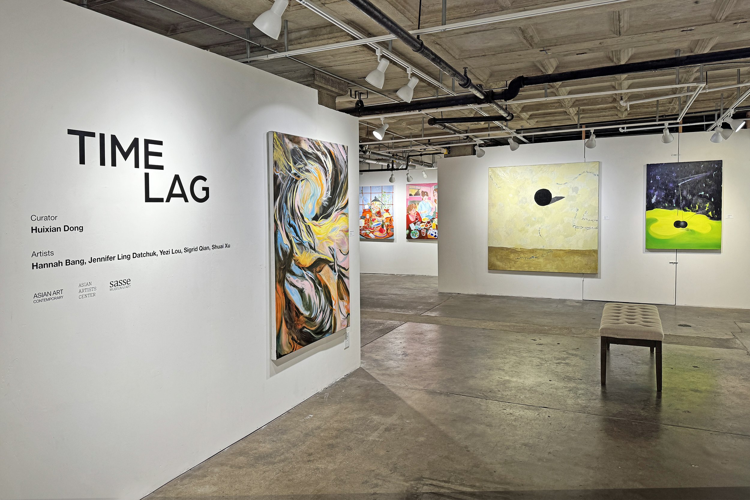 View of an art gallery with several colorful paintings on white walls, track lighting on a concrete ceiling, and a beige cushioned bench in the foreground.