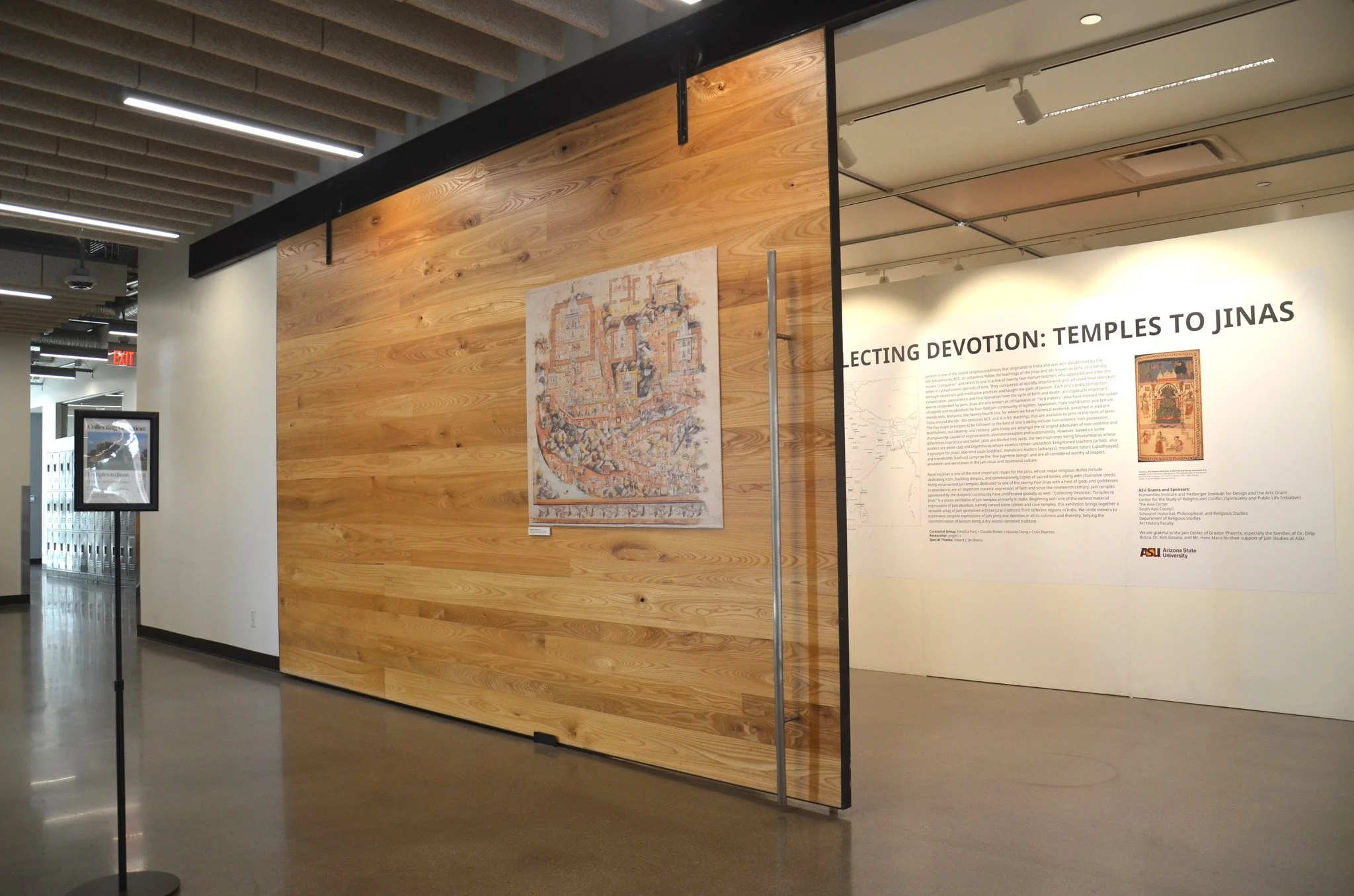 Exhibit display at a museum with wooden panel walls, featuring a map, artwork, and informational text about temples to Jinas, with a sign from Arizona State University.