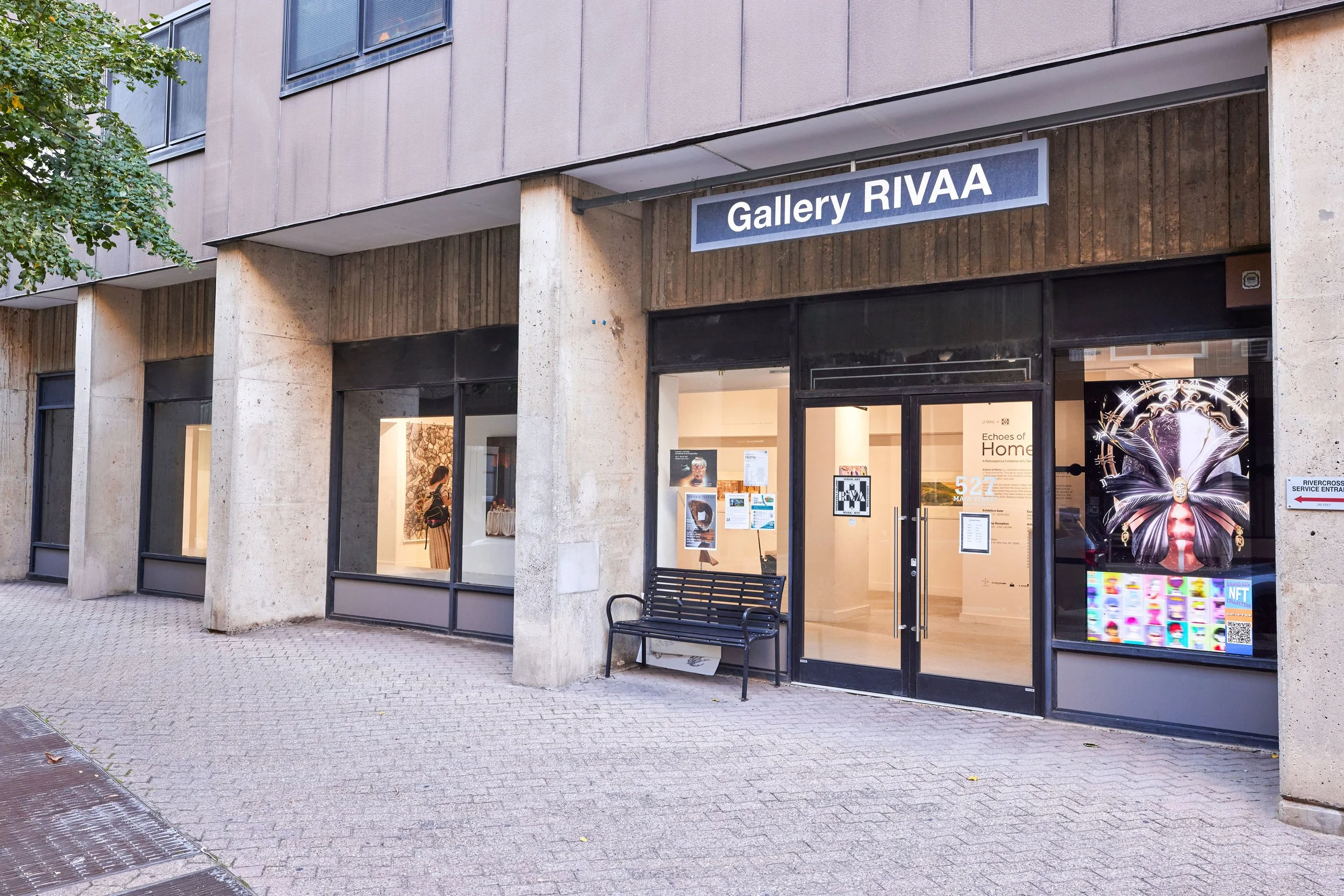 Exterior view of Gallery RIVAA art gallery with large glass windows, an entrance door, a bench outside, and artwork visible through the windows.
