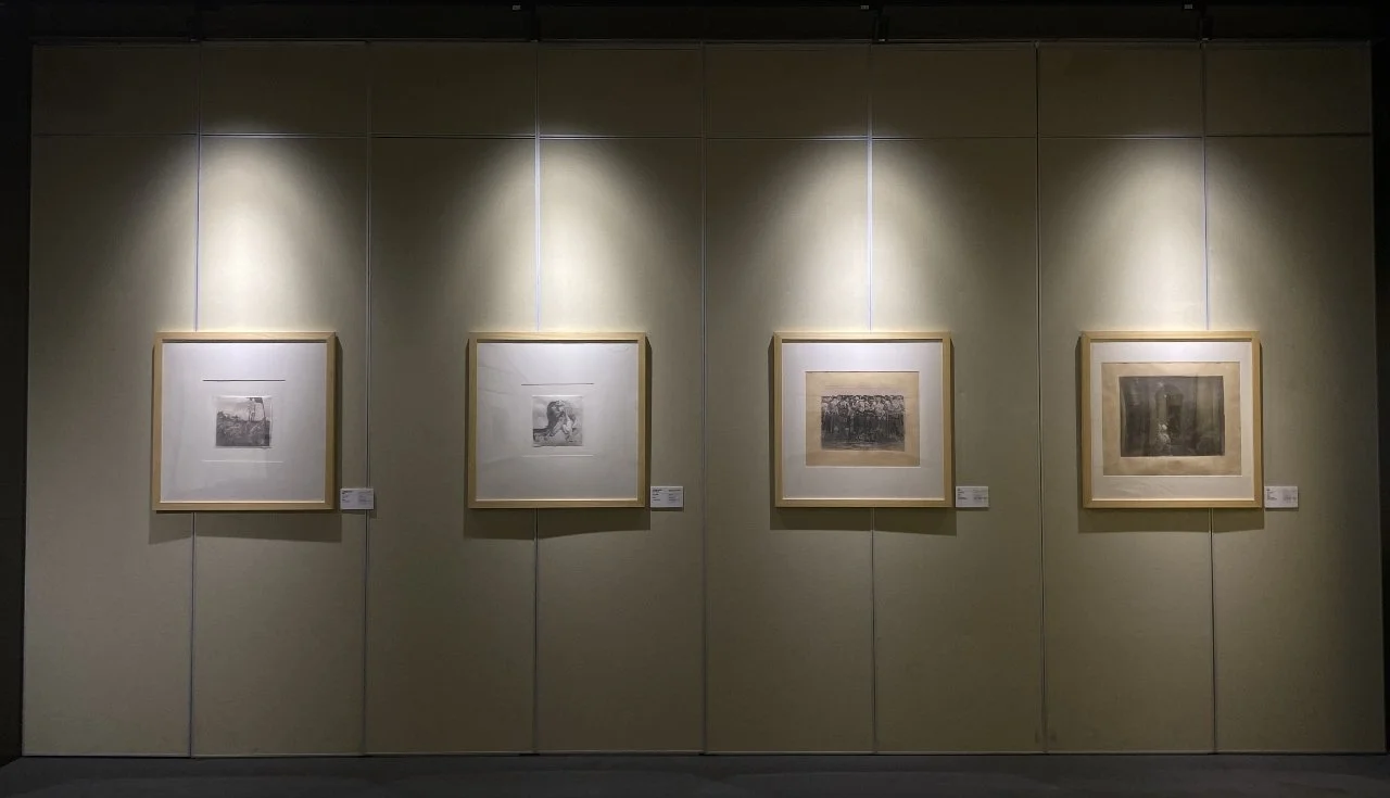 Four framed black-and-white photographs are hung on a beige gallery wall, each illuminated by a spotlight from above.