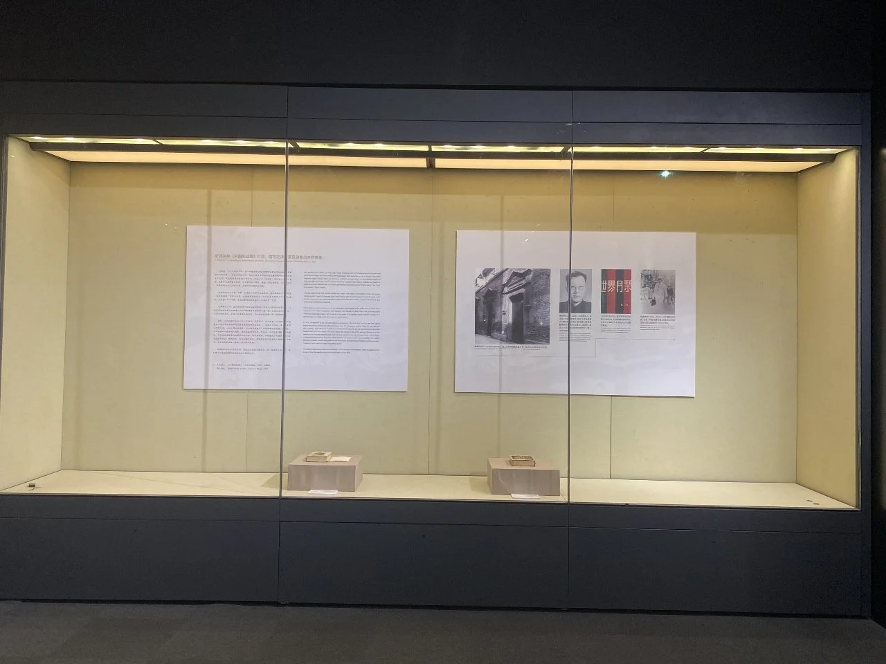 Display case with documents and photographs on interior walls, with two small objects on the bottom shelf.