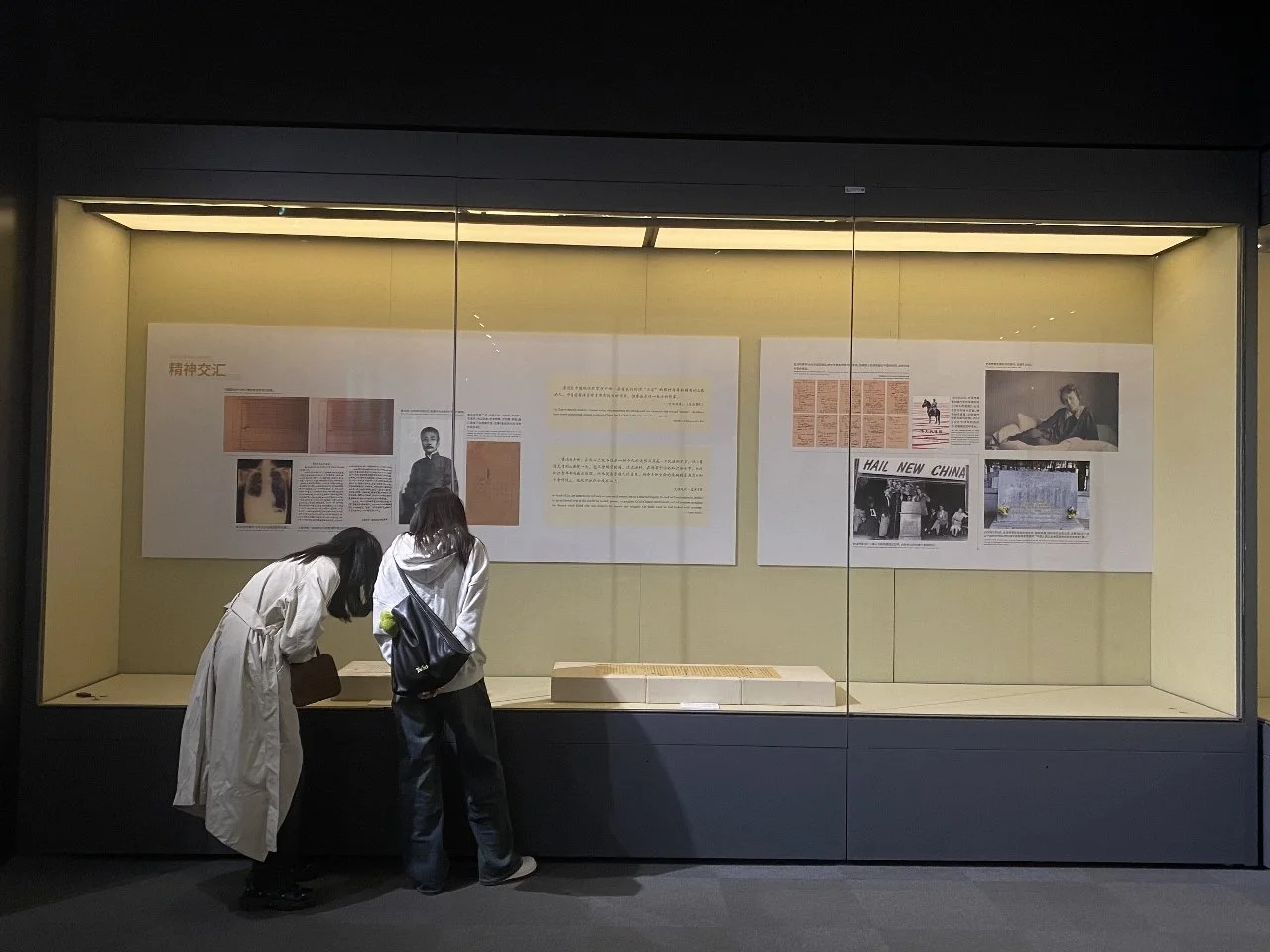 Two women are looking at an exhibition display case with text and historical photos inside a museum.