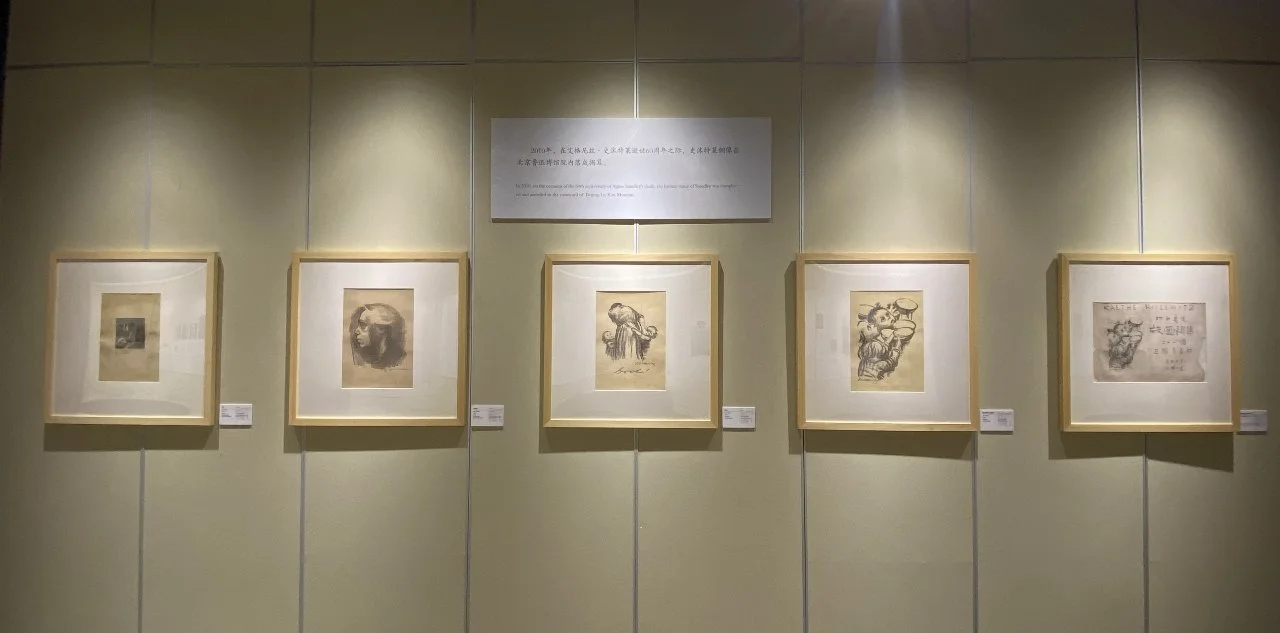 Exhibition wall with five framed sketches or artworks illuminated by wall-mounted lights, and a plaque with text above them.