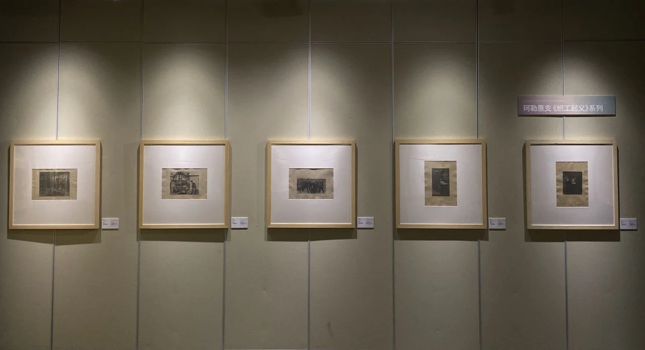 Five framed art pieces hanging on a light-colored gallery wall illuminated by spotlights.