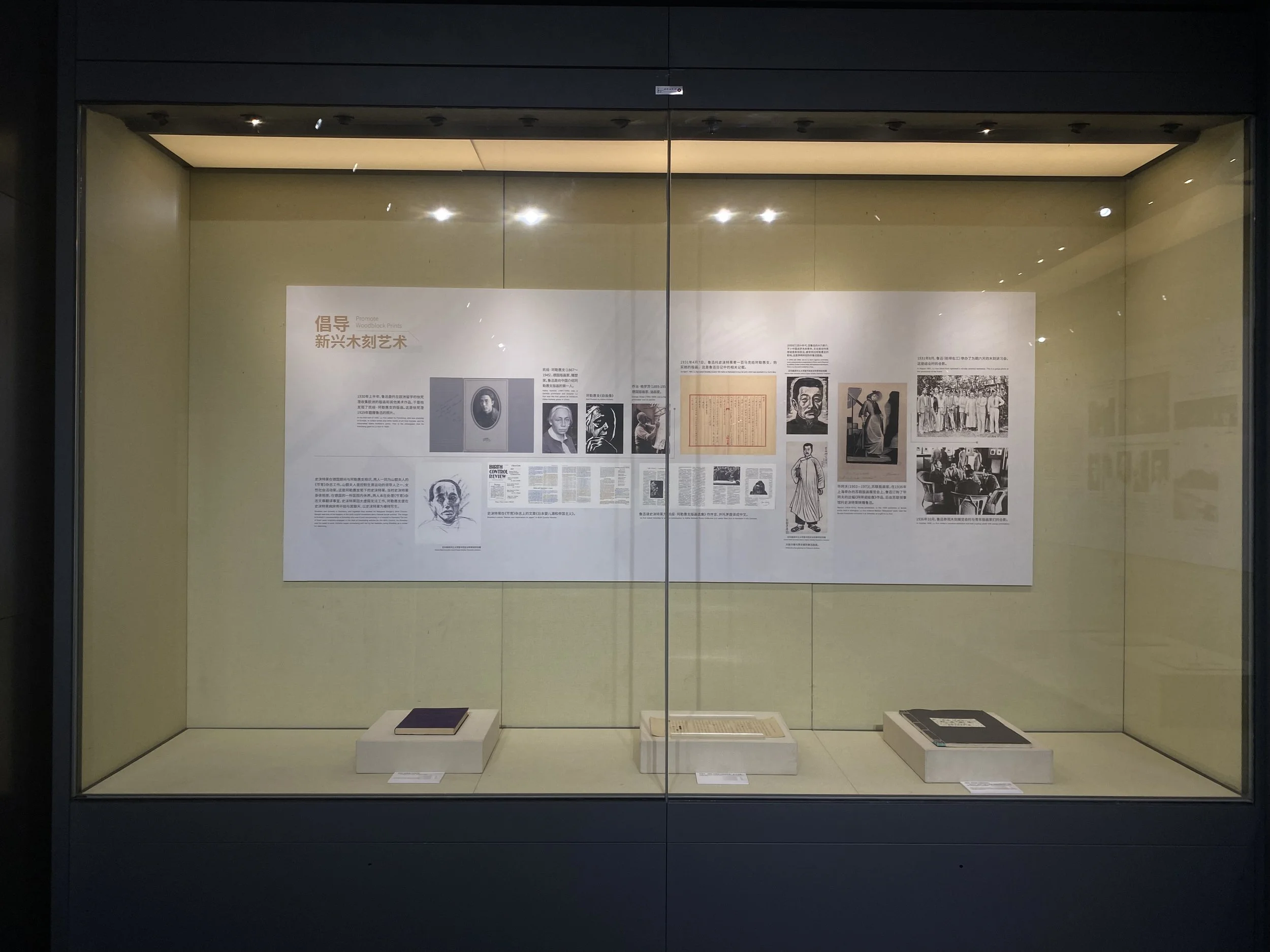 A museum display case with informational posters, black and white photos of individuals, and artifacts, including a framed document and a portrait of an elderly person.