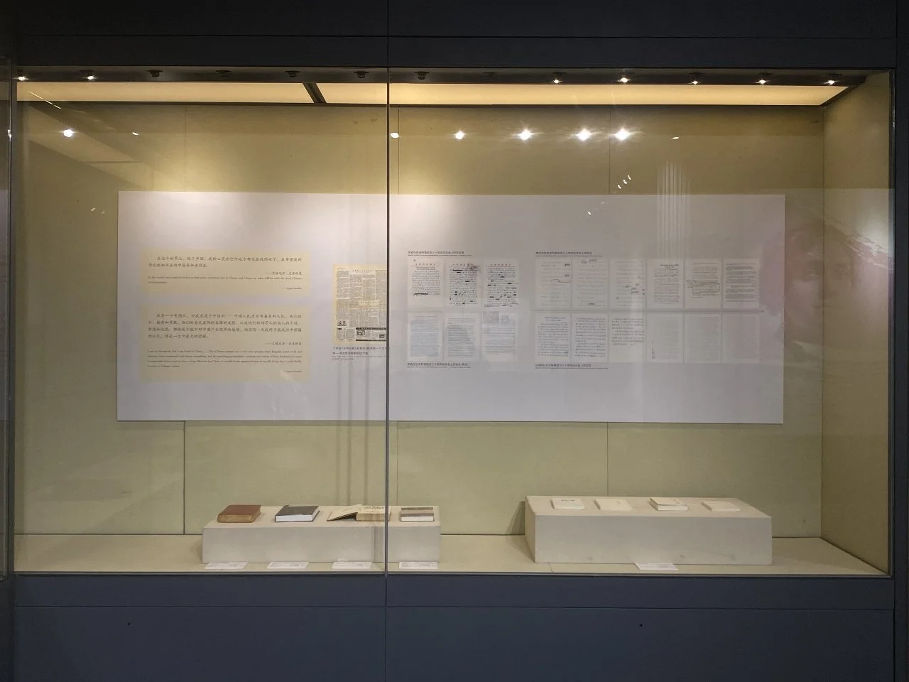 Museum display case with documents and books, showcasing printed materials on a white background, with some items arranged on platforms inside the case.