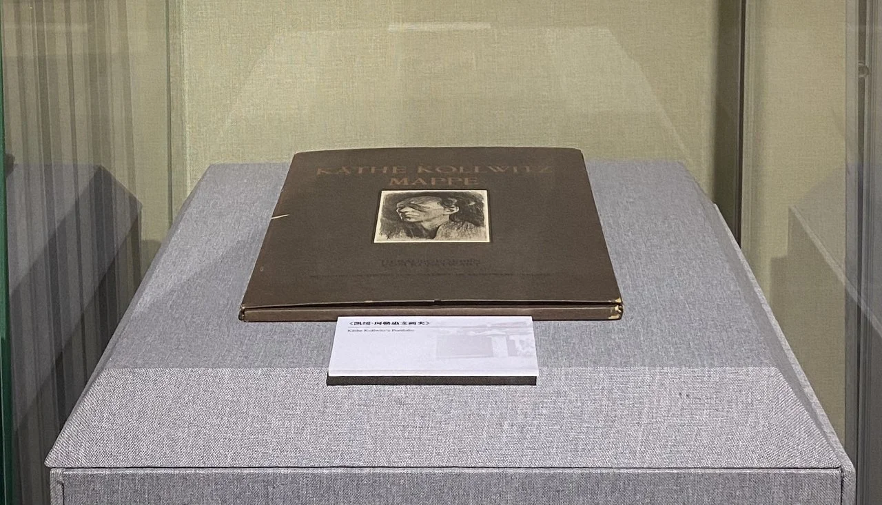 A book titled 'Kate Kollwitz: Mappé' displayed in a glass case on a gray fabric-covered stand, with a small label in front of it.