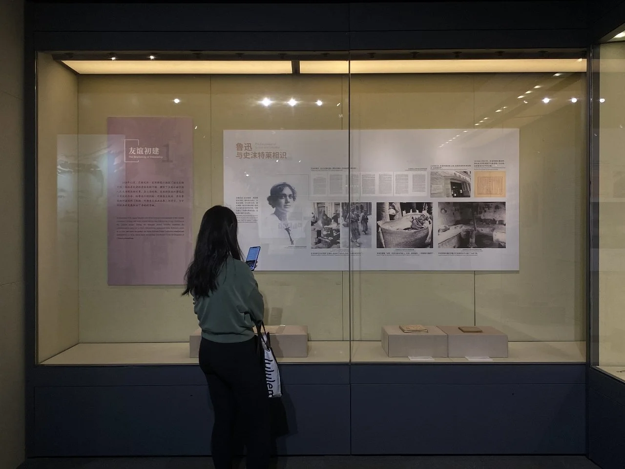 A woman with long black hair, wearing a gray top and black pants, is standing in front of a museum display case, looking at informational panels and photographs behind glass.