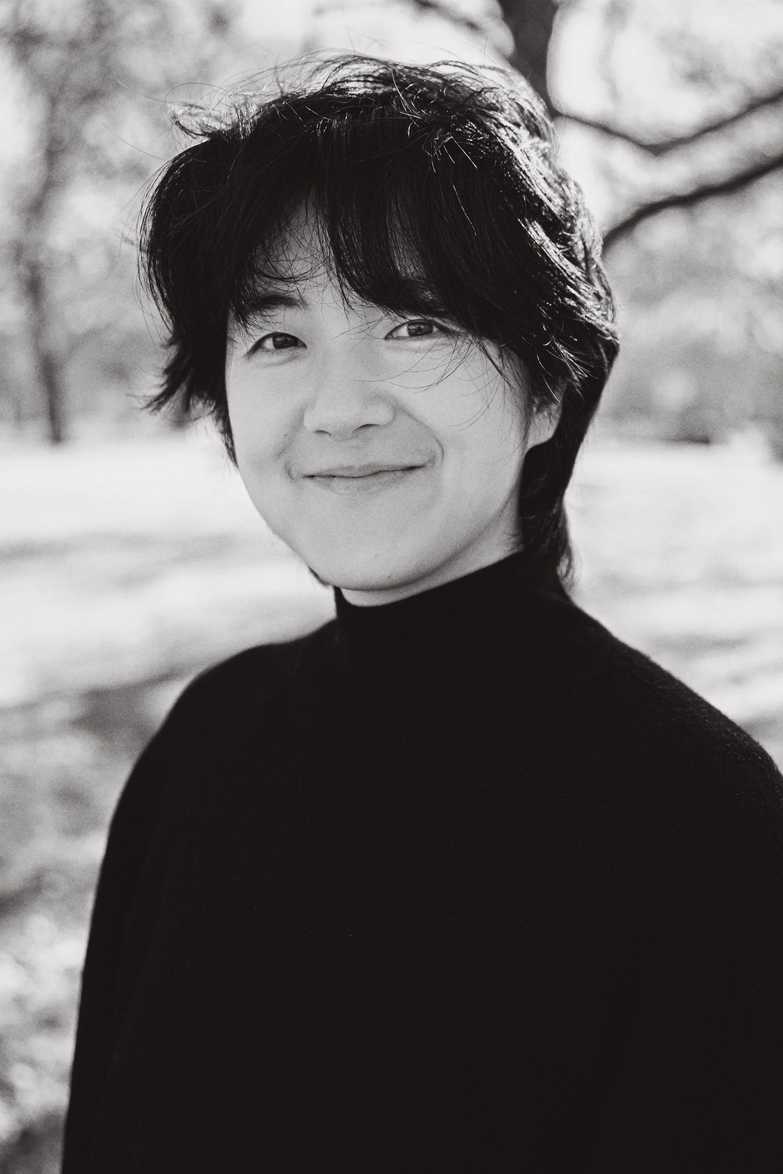Black and white portrait of a woman with short wavy hair, smiling softly and looking slightly to the left. She is wearing a dark turtleneck sweater, and the background appears to be outdoors with blurred trees.