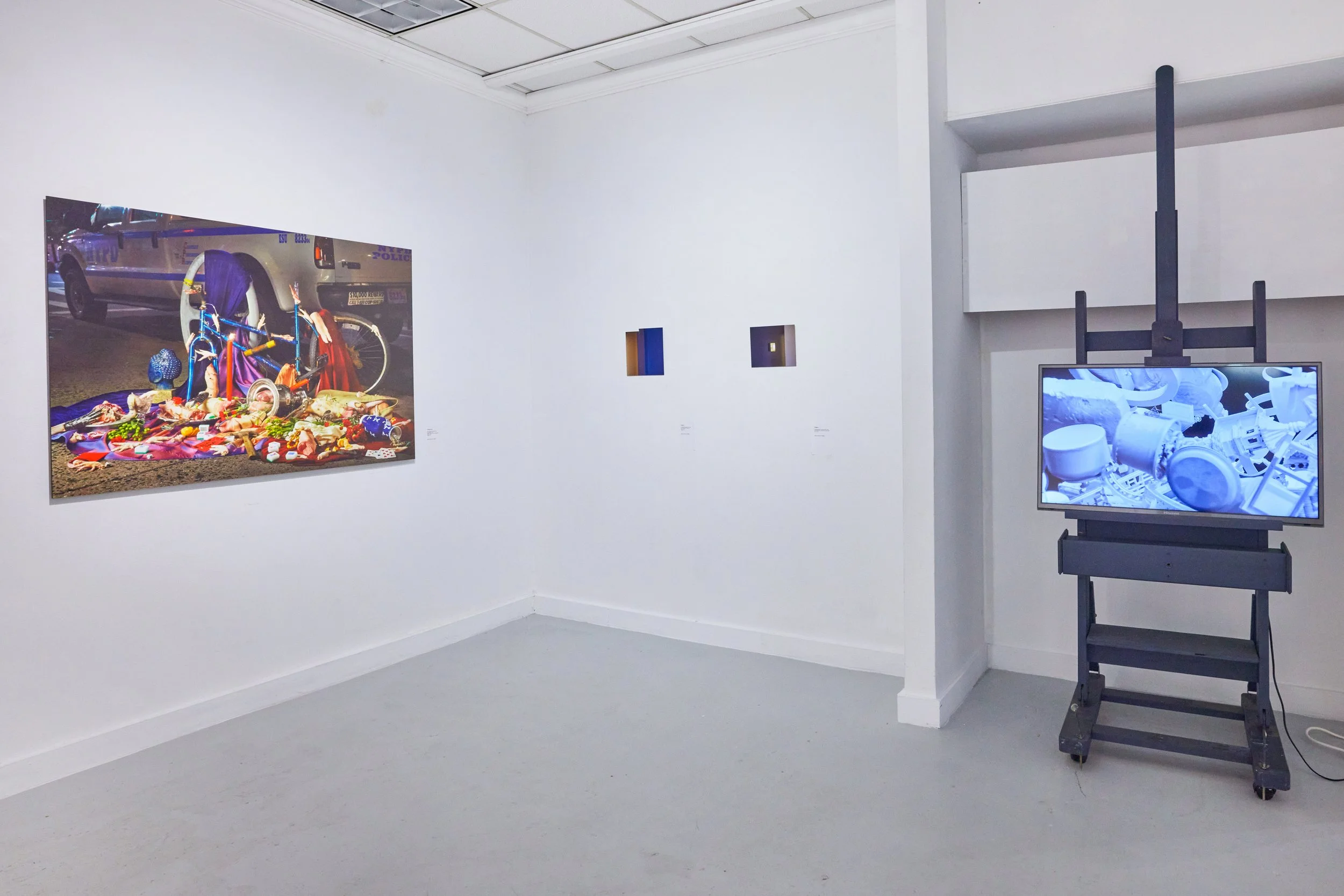 An art gallery with white walls displaying paintings and digital screens. One painting depicts a scene of vandalized police cars and overturned bicycles with various objects and debris. The other screen shows abstract imagery in blue and white tones.