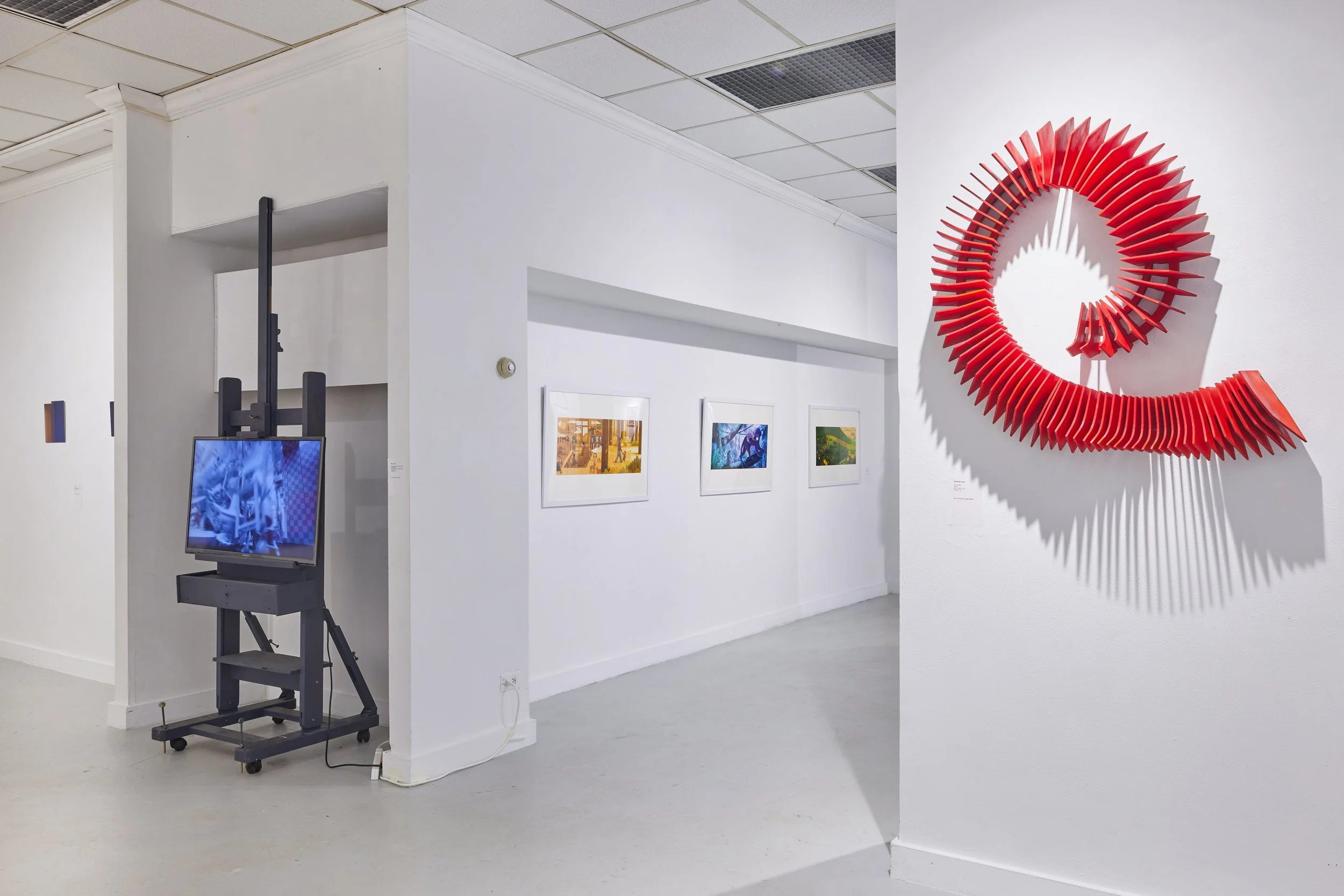An art gallery with white walls and ceiling. There are framed artworks on the wall, and a large red circular sculpture with radiating spokes is mounted on one wall. A monitor on a black stand is in the corner, showing a blue image with what appears t