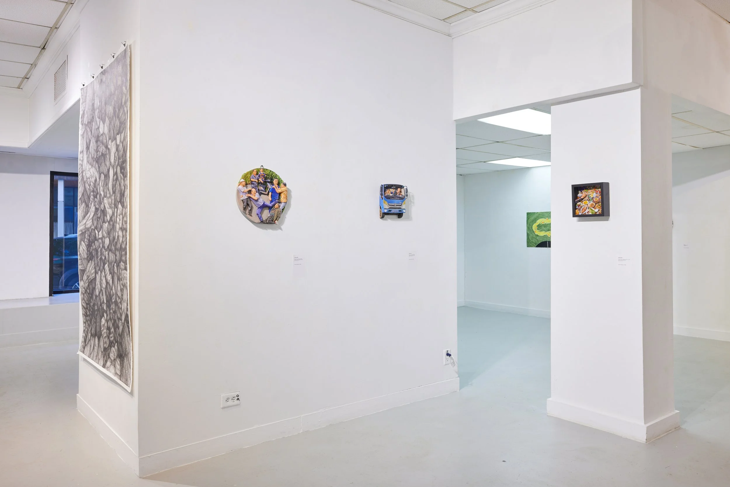 Photo of an art gallery interior with various artworks on white walls, including a circular artwork of people, a small print of food, and a green painting with yellow design. The space has white walls, a ceiling with square tiles, and a corner with a