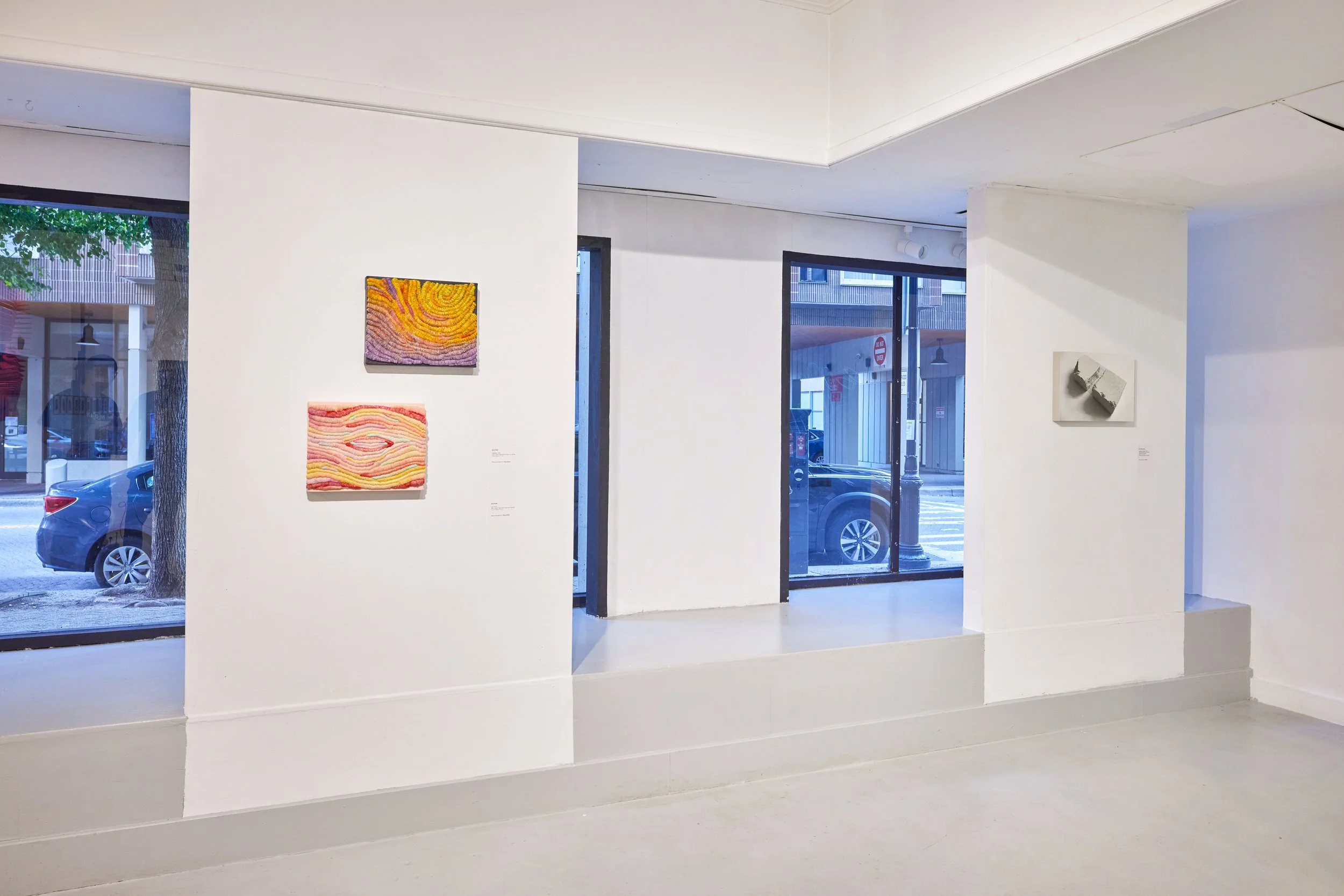 Interior view of an art gallery with white walls showcasing colorful textured artwork. Large glass windows reveal parked cars and a street scene outside.