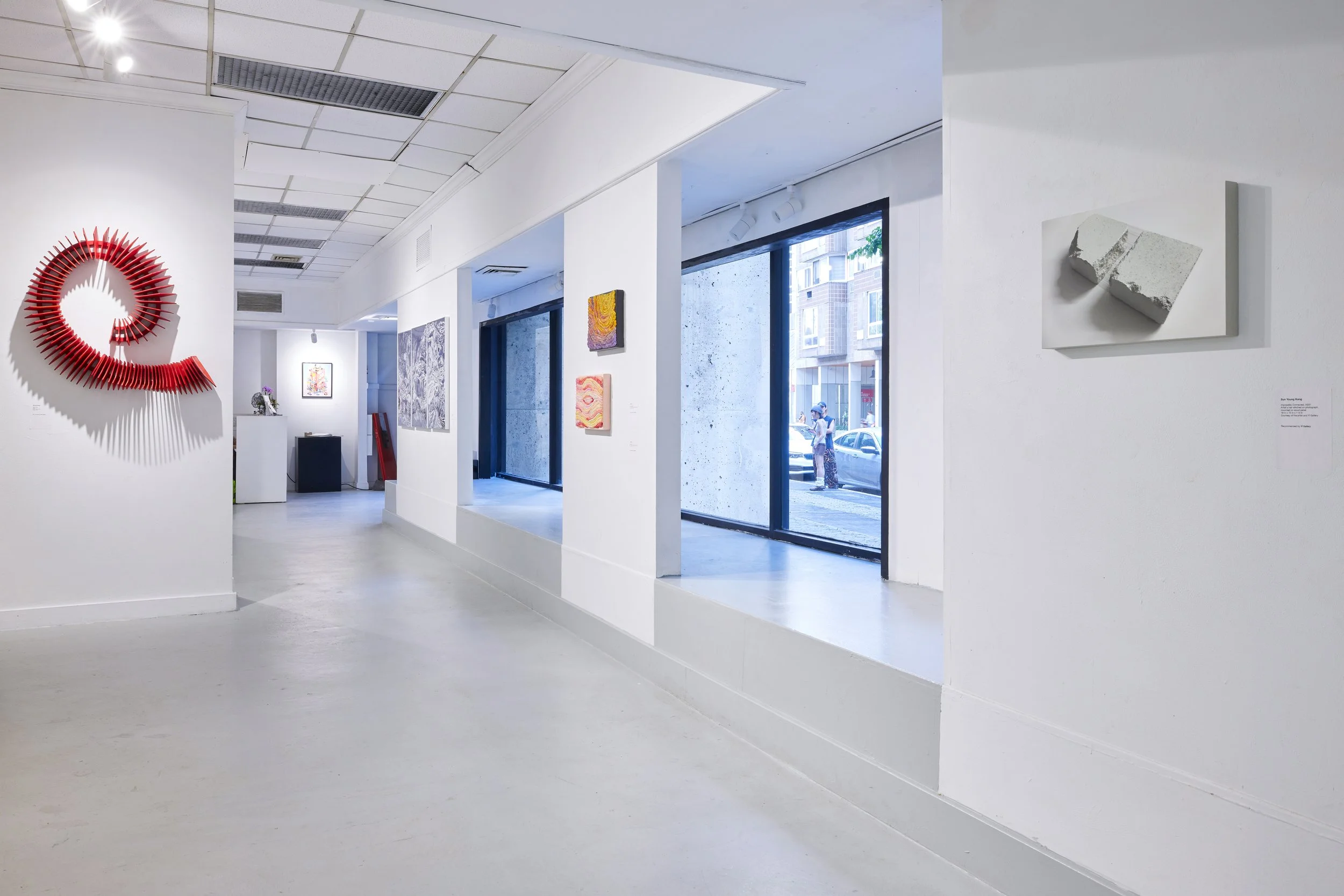 Interior view of an art gallery with white walls, several colorful abstract paintings, a large red circular sculpture on the wall, and large windows showing an urban street outside.