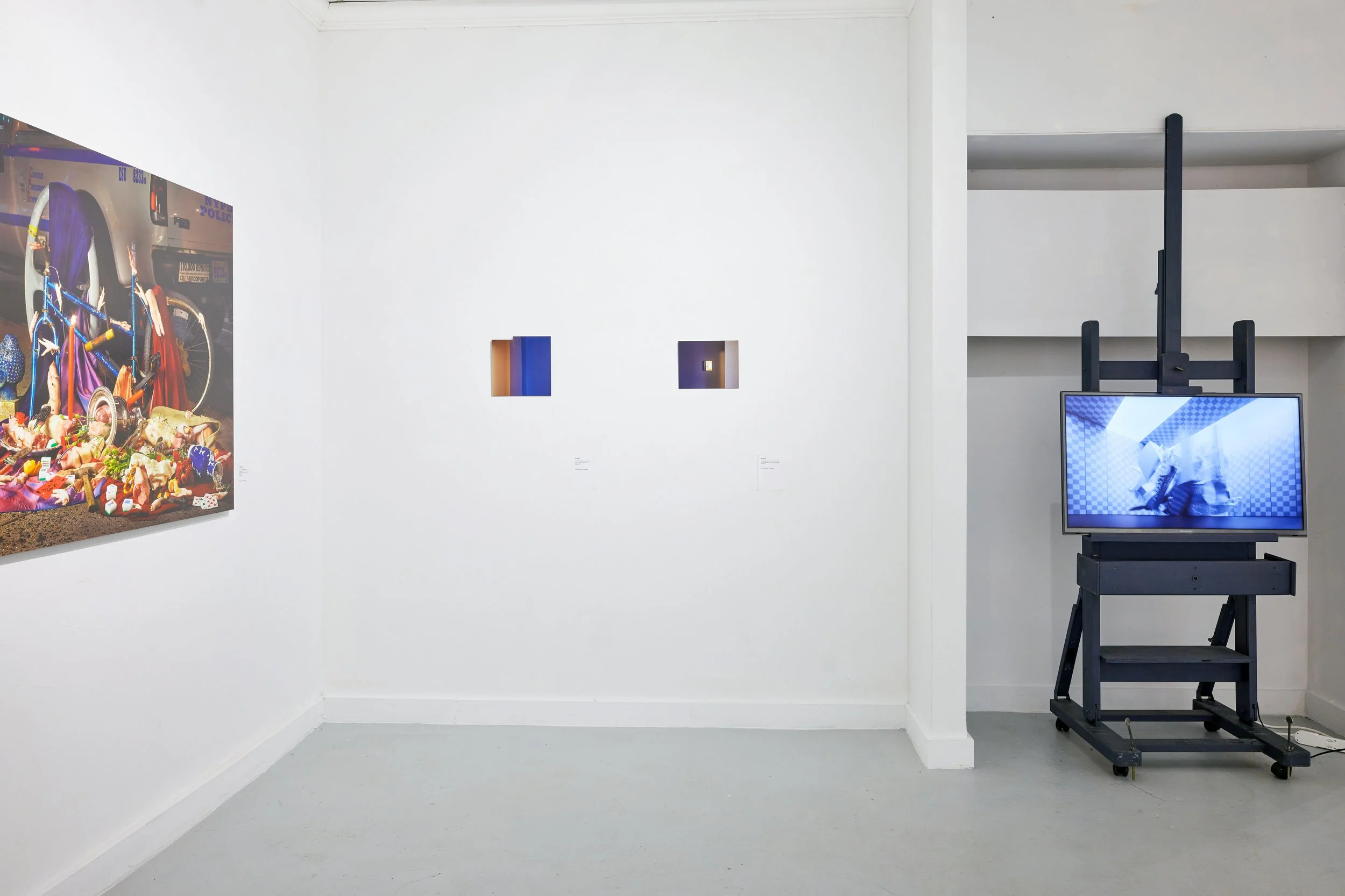 Art gallery with a white wall featuring a large colorful painting on the left, two small square artworks in the middle, and a digital screen on a black stand on the right. The gallery space has gray flooring and minimal decor.