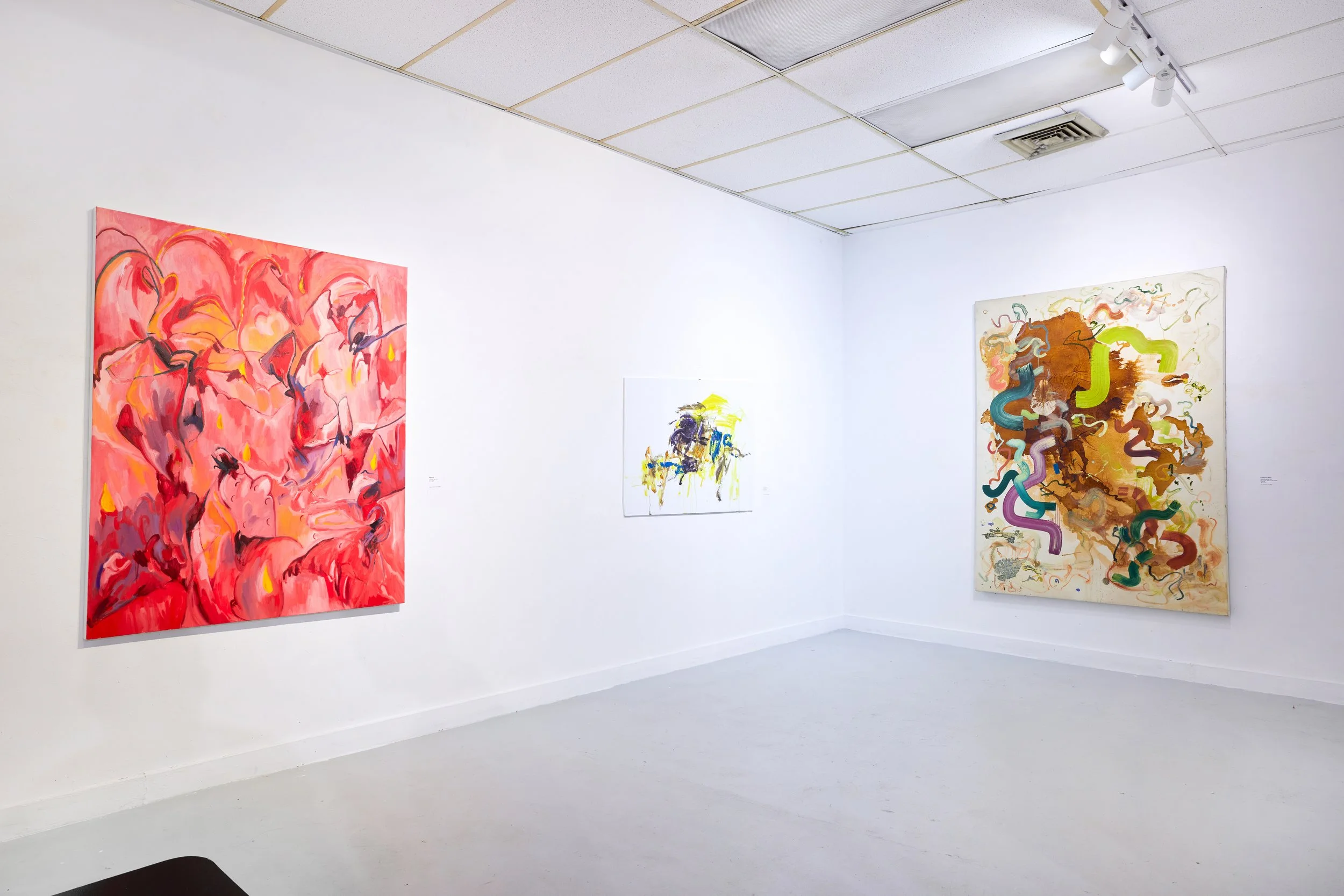 An art gallery with white walls displaying colorful abstract paintings, including a pink and red piece on the left and a multicolored piece on the right, with a white ceiling and track lighting.