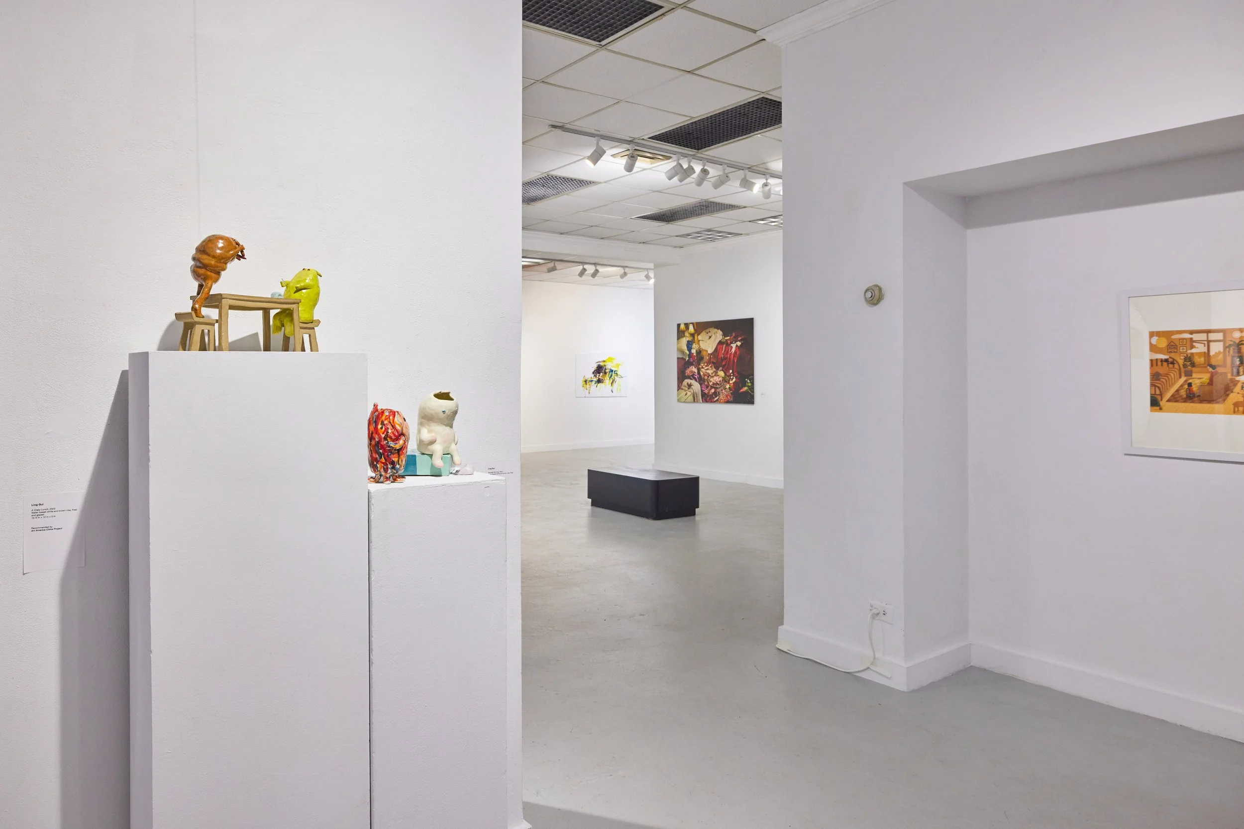 Contemporary art gallery displaying colorful sculptures on white pedestals and paintings on white walls, with a black bench in the middle of the room.