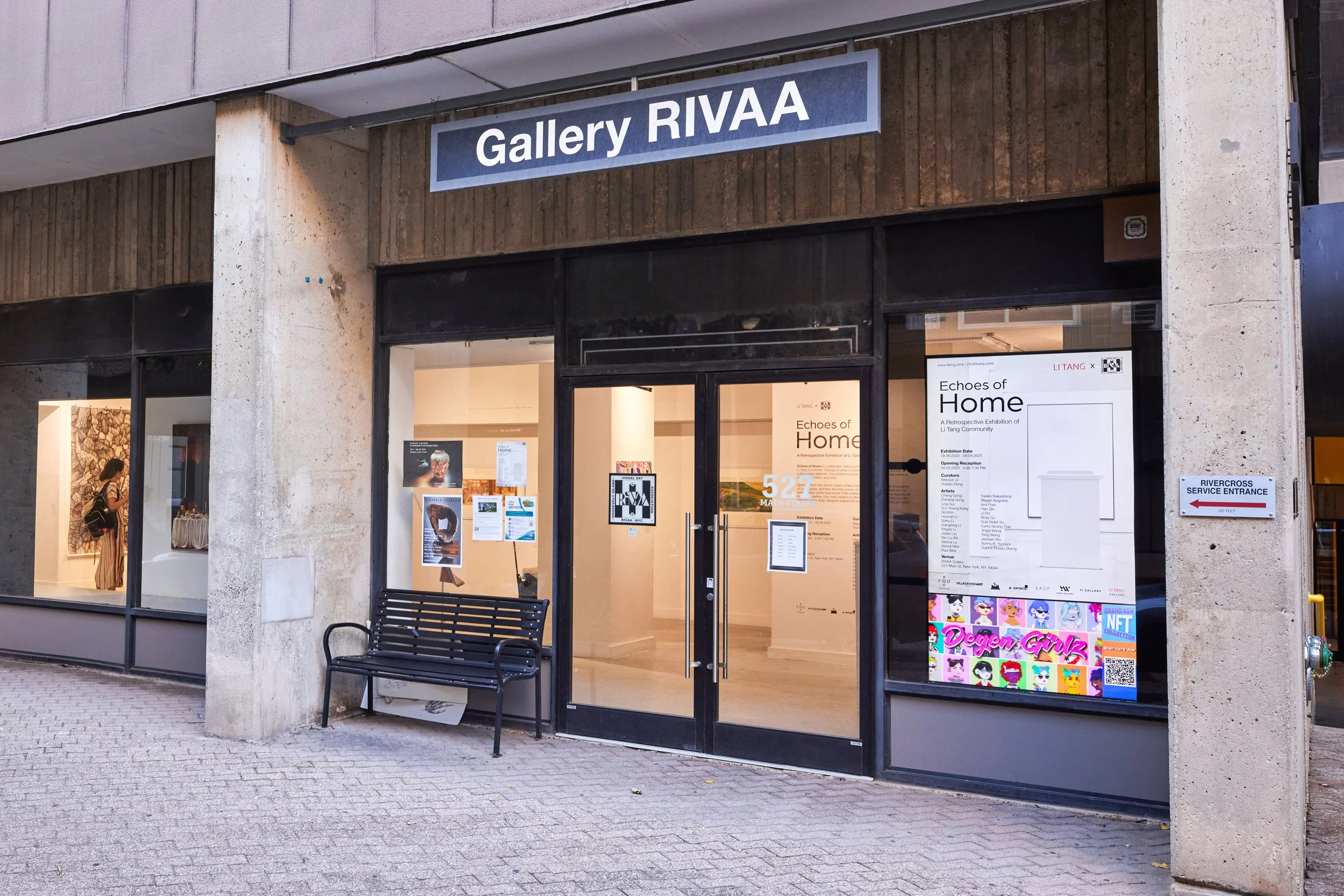 The exterior of an art gallery building named 'Gallery RIVAA,' with large glass doors displaying posters and advertisements for art events, and a black bench outside.
