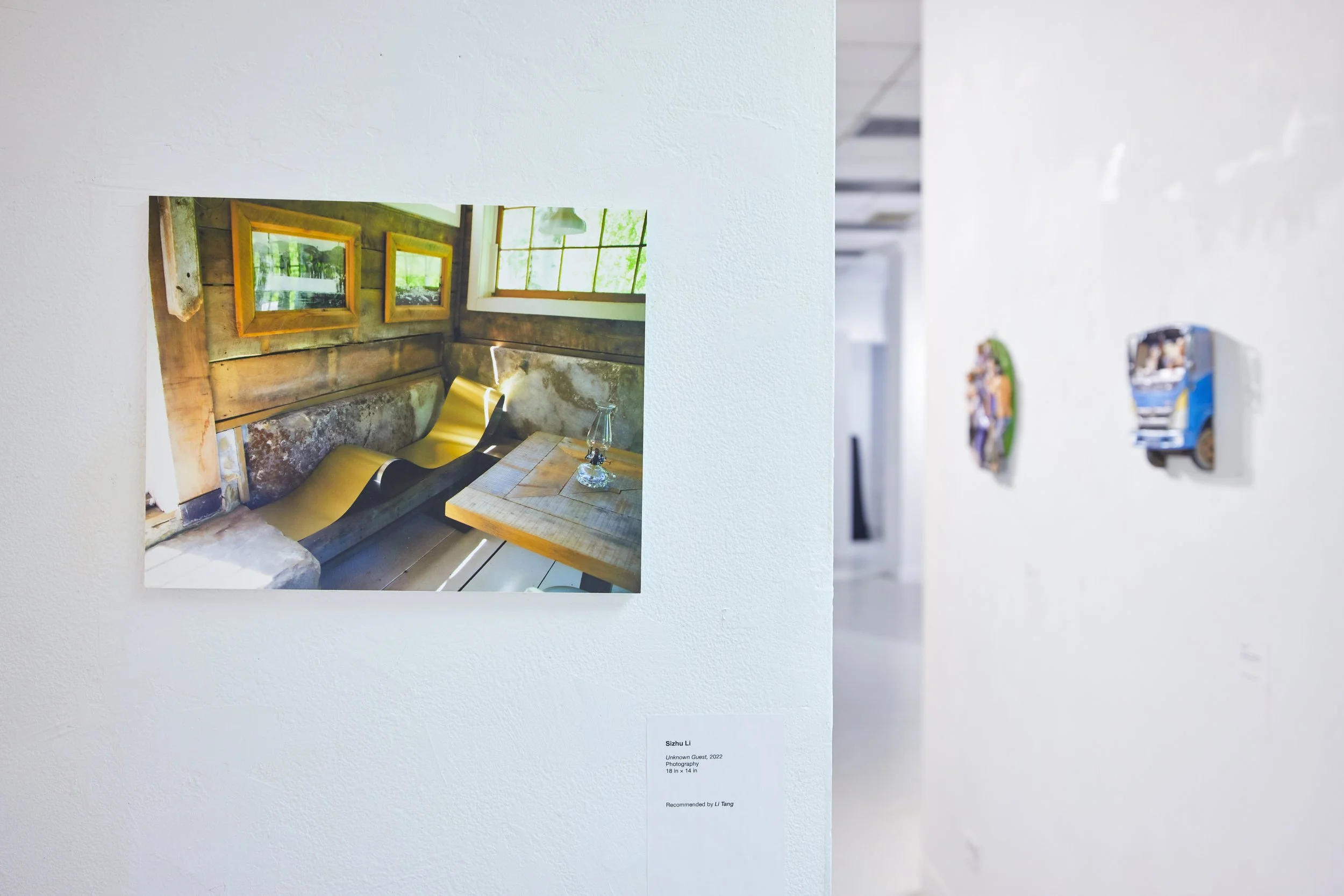 Photograph of an interior with rustic wooden walls, three small windows, and a yellow bathtub with a bottle and mirror on the wooden surface. Exhibition label underneath. Bright natural light coming through the windows.