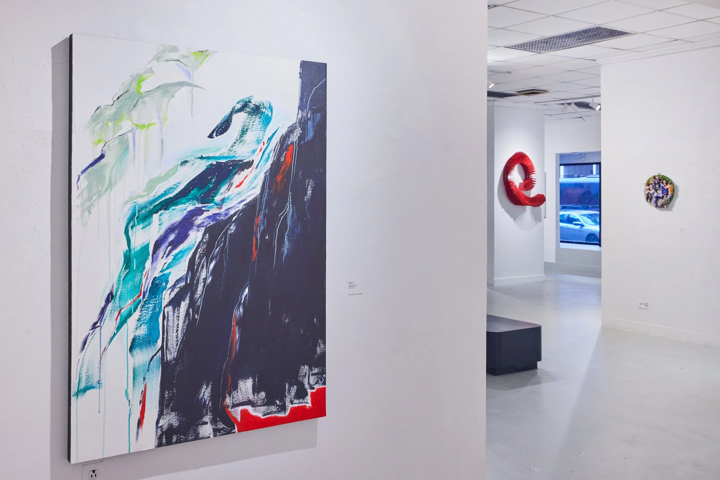 Abstract colorful painting displayed on a white gallery wall, with a red sculpture and a round artwork visible in the background inside a modern art gallery.