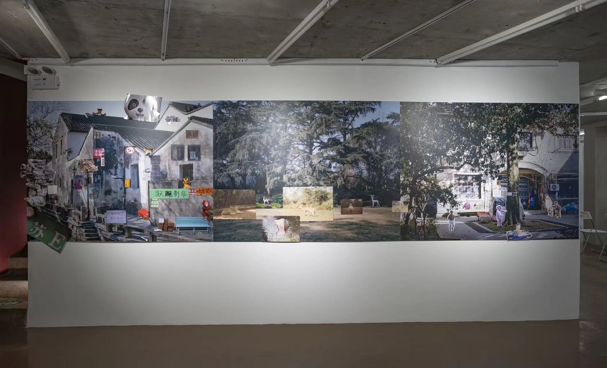 A mural on a white wall features a collage of outdoor scenes including trees, a house, and people, with small cutouts and illustrations, in an indoor setting with a concrete ceiling.
