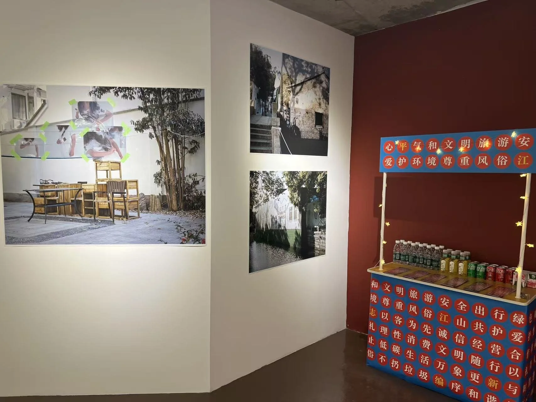 Two wall-mounted photographs of outdoor scenes, one featuring a patio with chairs and a tree, the other showing steps and a building, displayed on a white wall. To the right, a table with bottled drinks and a red backdrop with text in Chinese charact