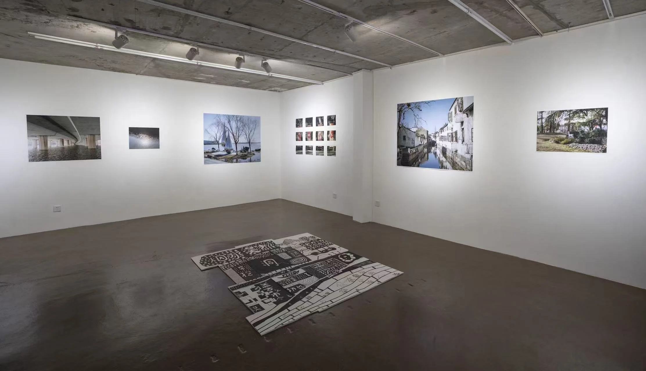 Art gallery with framed photographs of scenes including waterways, trees, and buildings on white walls. Carpet with black and white abstract and text designs on a concrete floor.