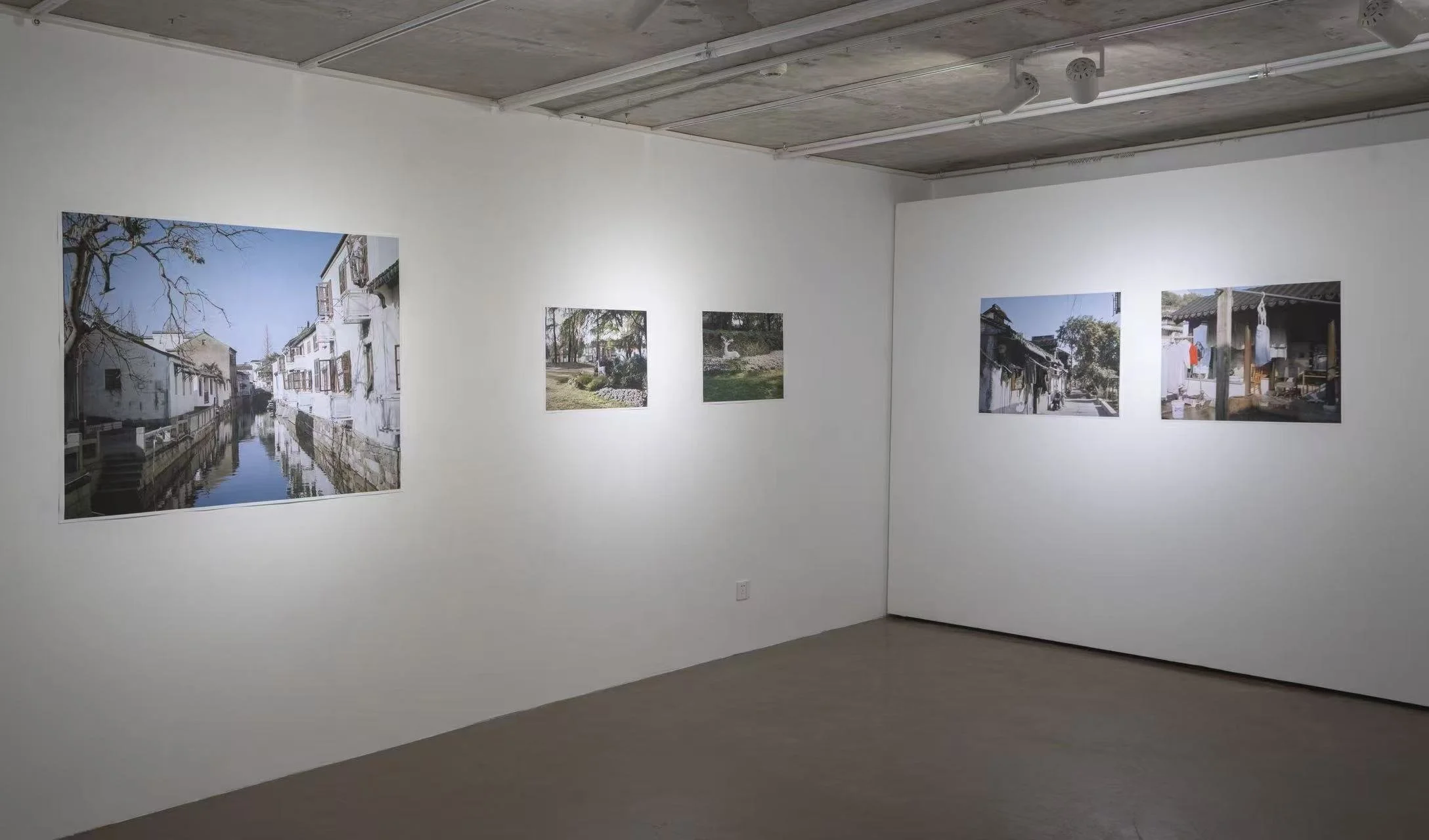 Gallery interior with white walls displaying framed landscape photographs of outdoor scenes, including buildings, trees, and water, in an art exhibition setting.