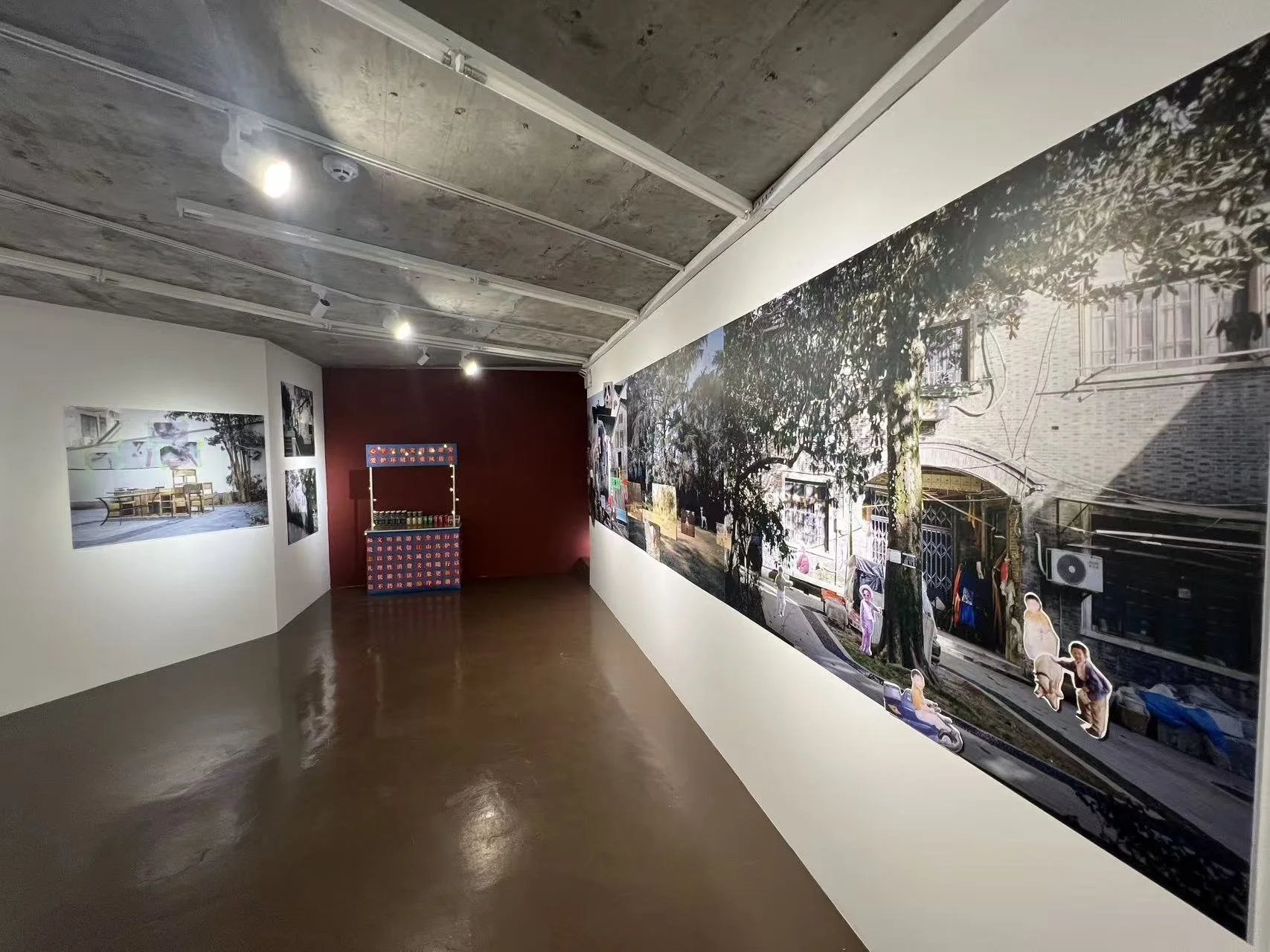 An art gallery with large photographs on the walls, a red wall at the end, spotlights on the ceiling, a wooden floor, and a small display stand.