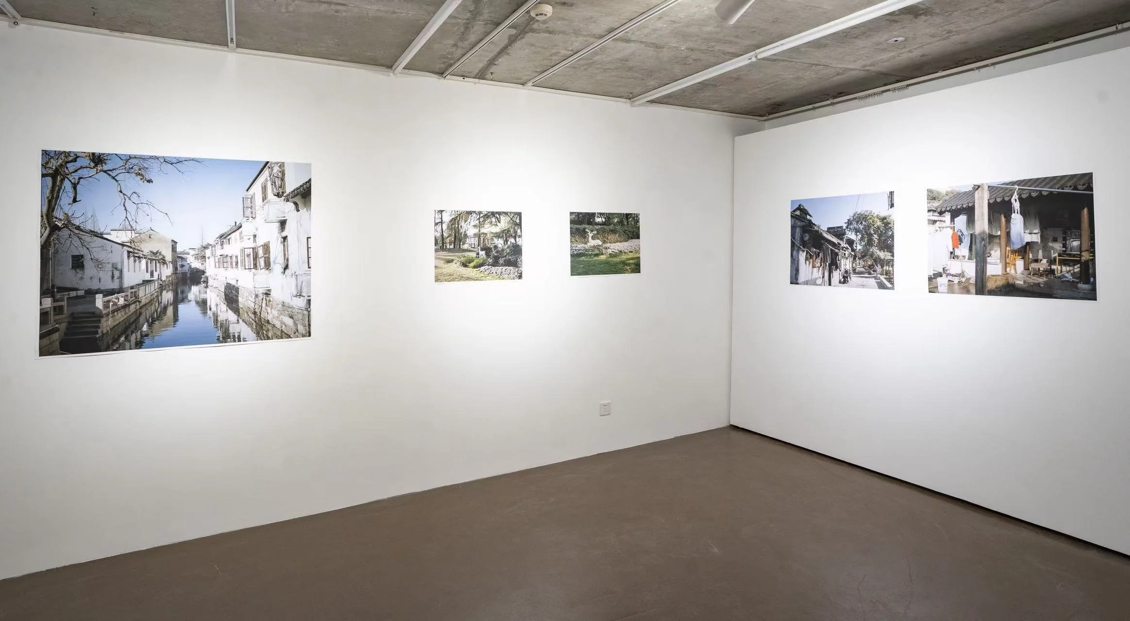Gallery wall with multiple photographs of outdoor scenes, including a canal, trees, and buildings, in an art exhibit space with white walls and a beige floor.