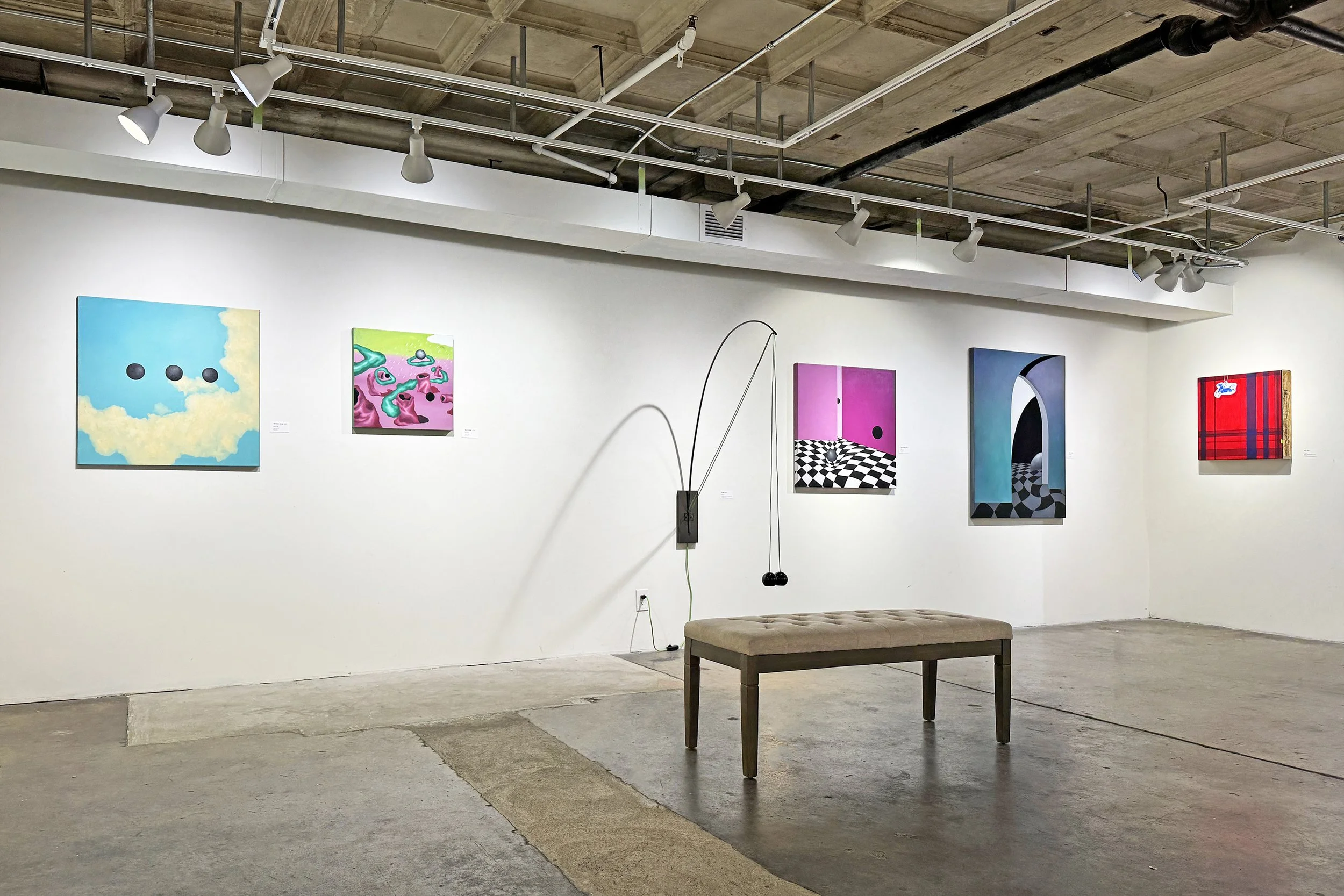 An art gallery with white walls displaying colorful paintings; the ceiling features exposed pipes and track lighting; a cushioned bench is in the foreground.