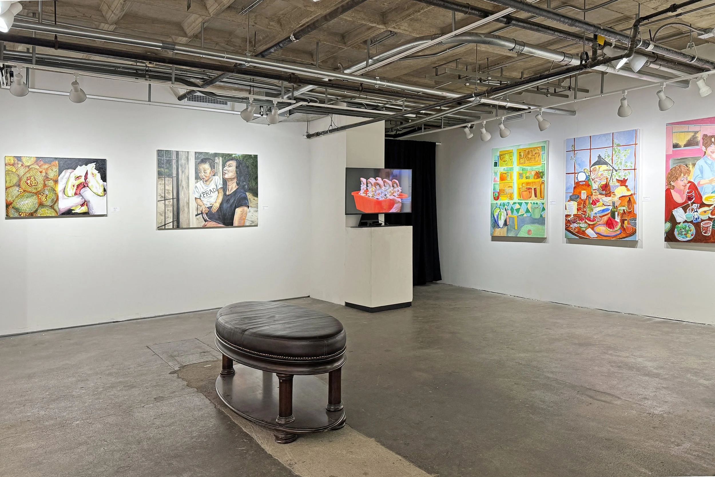 Gallery wall with multiple colorful paintings in an art exhibition space with a concrete floor and ceiling. Furniture includes a round leather seat in the foreground.