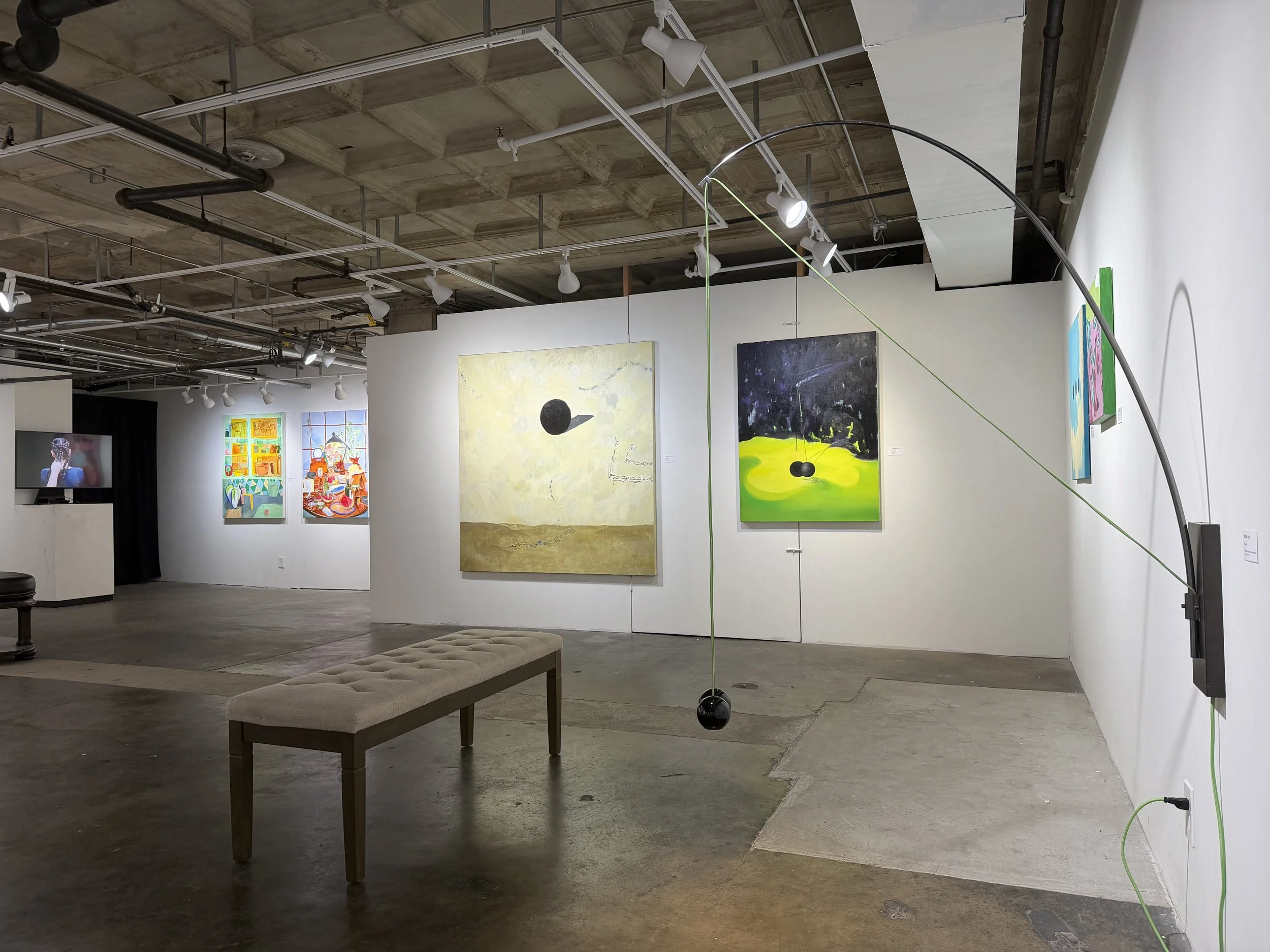 Image of an art gallery with paintings hanging on white walls, ceiling lighting, a bench in the foreground, and an art installation hanging from the ceiling consisting of black and green spheres connected by strings.