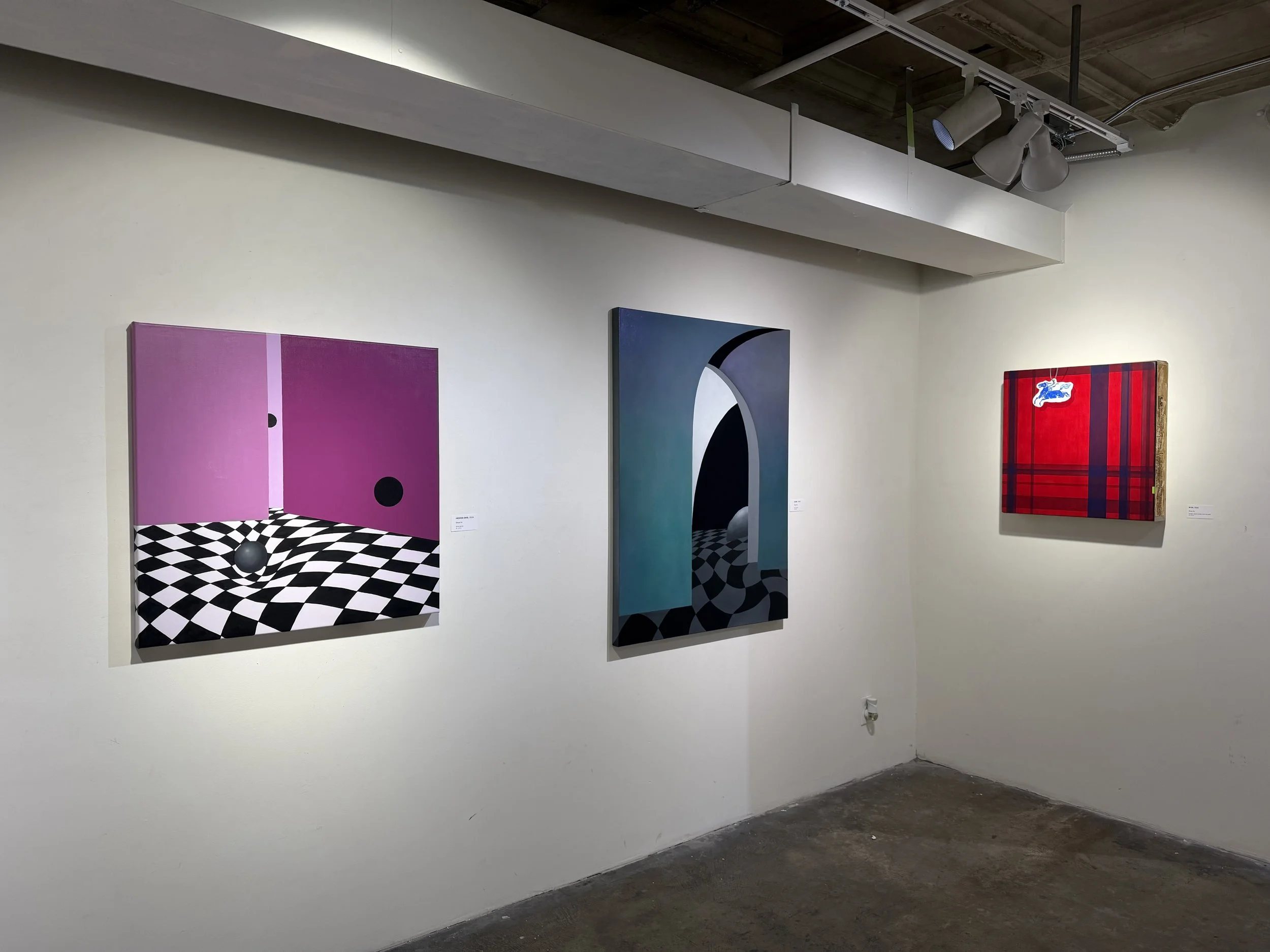 Three colorful abstract paintings displayed on a white gallery wall with spotlights and a concrete floor.