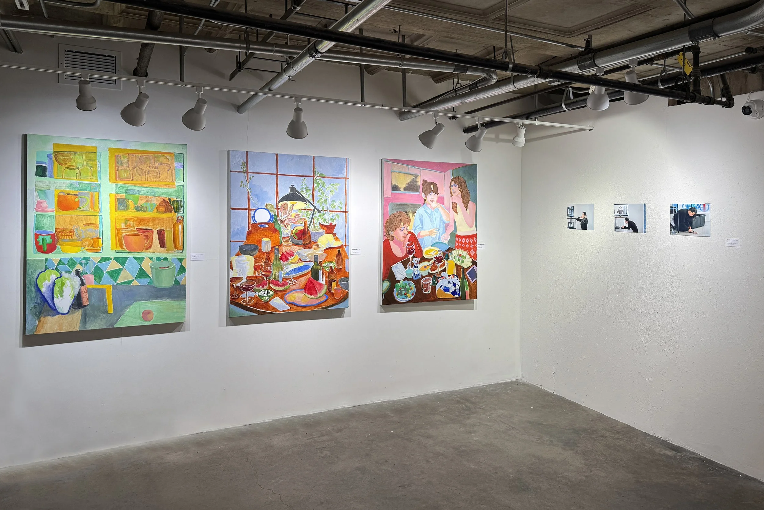 Three colorful paintings are displayed on a white gallery wall. The paintings depict vibrant still life and interior scenes, with various objects, plants, and people. The gallery has ceiling track lighting and visible industrial ceiling pipes.