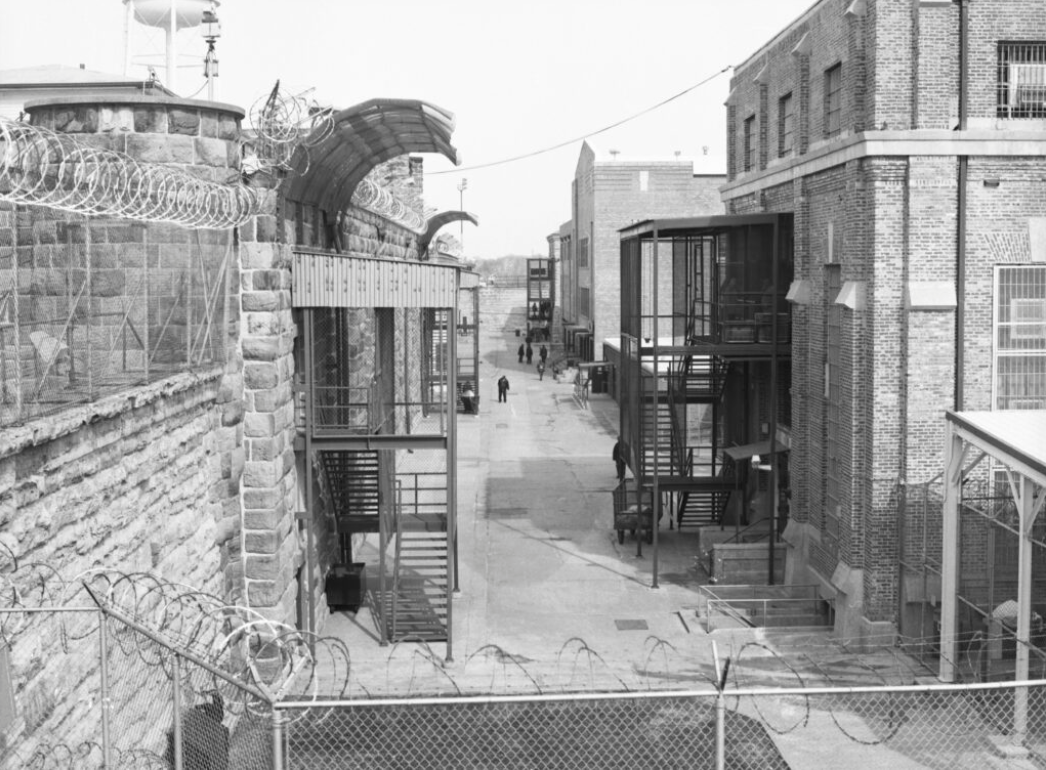 Historical prison yard with barbed wire, stone and brick walls, and metal staircases, viewed from behind a wire fence.
