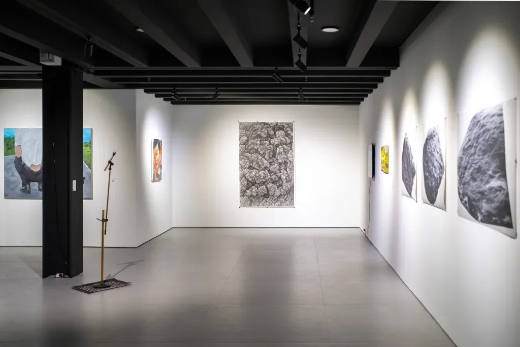 An art gallery with various paintings on white walls, including a large black-and-white textured piece in the center and several other artworks on the right, along with a sculpture and a mic stand on the left side.
