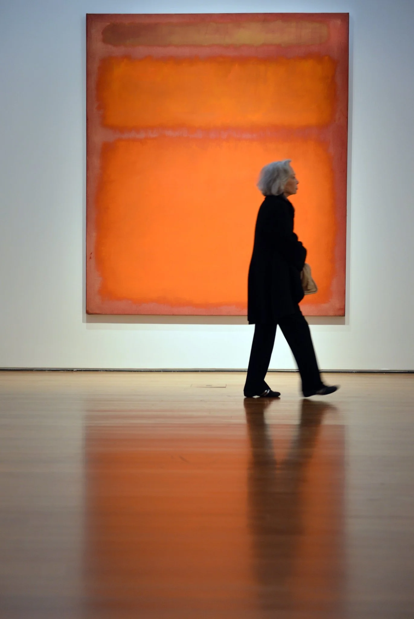 An elderly woman with gray hair, dressed in black, walking past a large abstract orange and red painting in an art gallery.