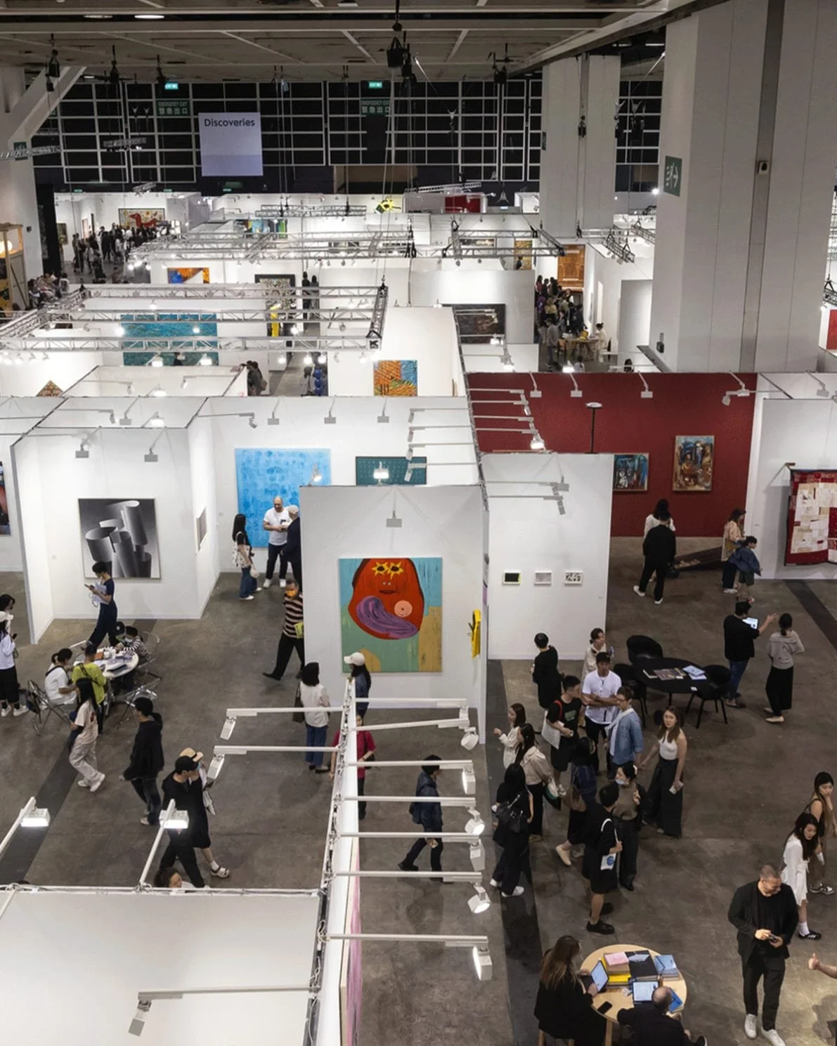 An indoor art exhibition with white display walls showcasing various paintings, visitors walking and viewing art, and some people sitting at tables, in a spacious modern hall with high ceilings.