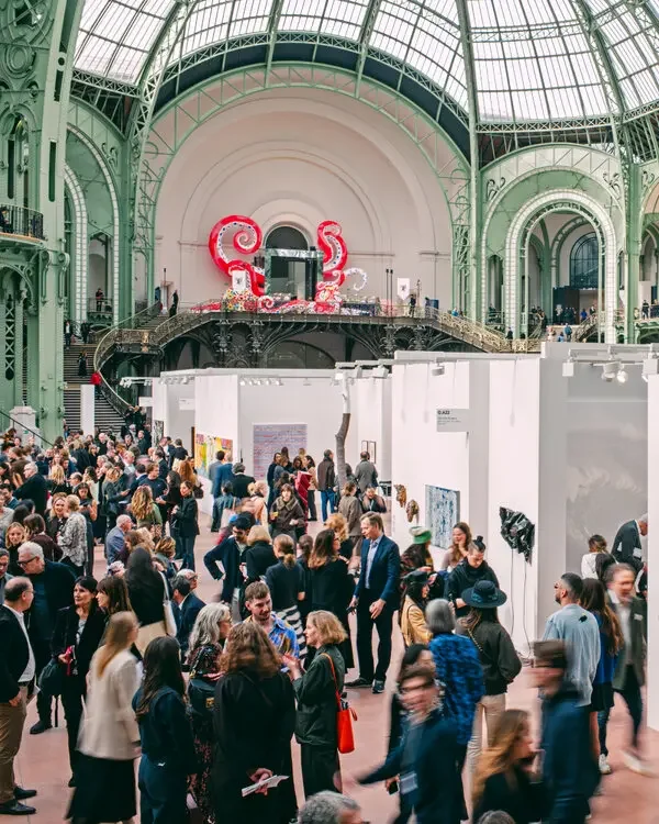 Crowd of people in an art gallery or exhibition hall with white partition walls displaying artwork, inside a historic building with a glass domed roof and green architectural details. An ornate staircase and colorful decorations are visible in the background.